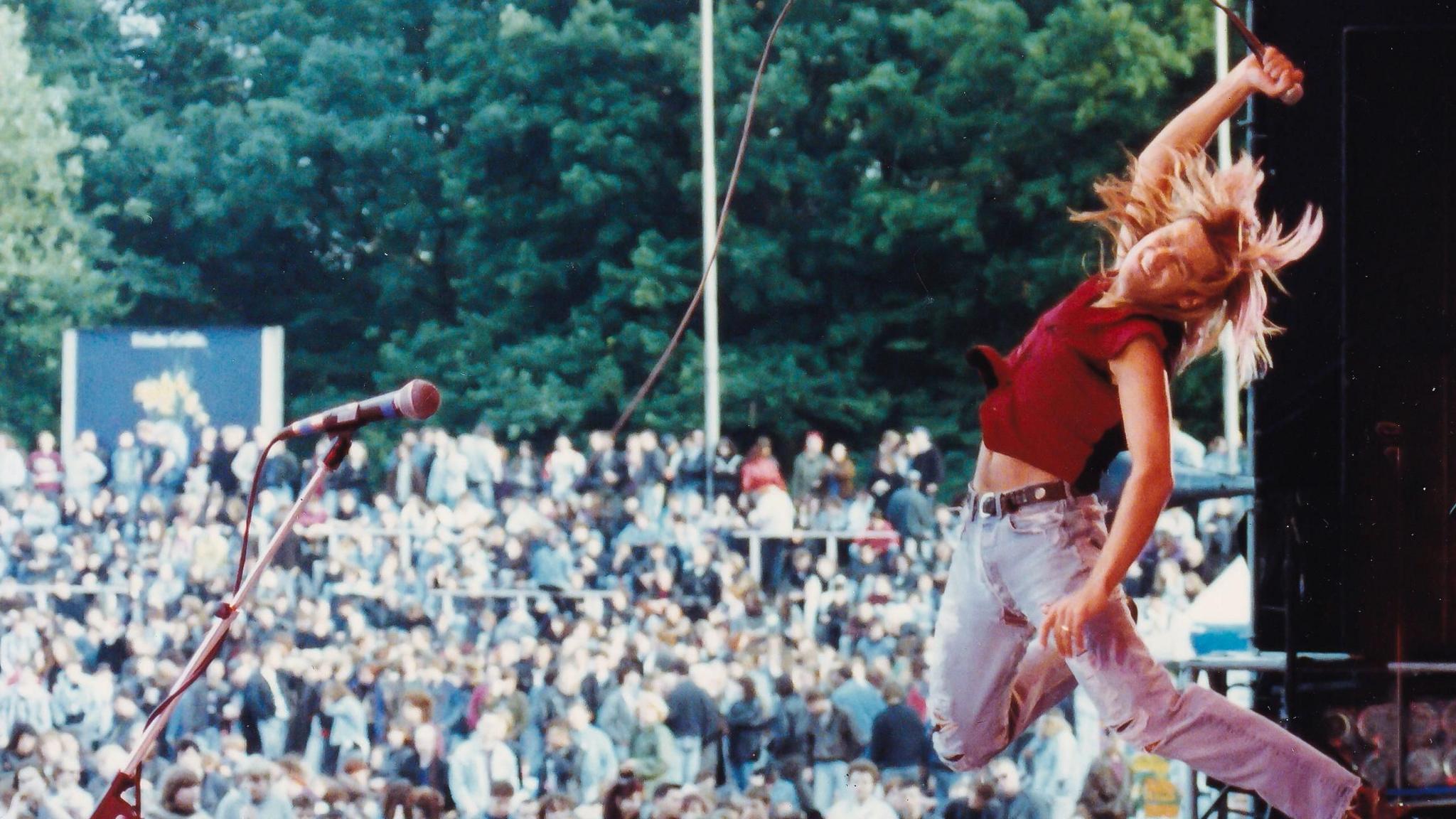 Mike Peters jumps in the air on stage holding a microphone up, with the crowd in the background. His long blonde hair flies around him. He is wearing a red shirt and ripped jeans.