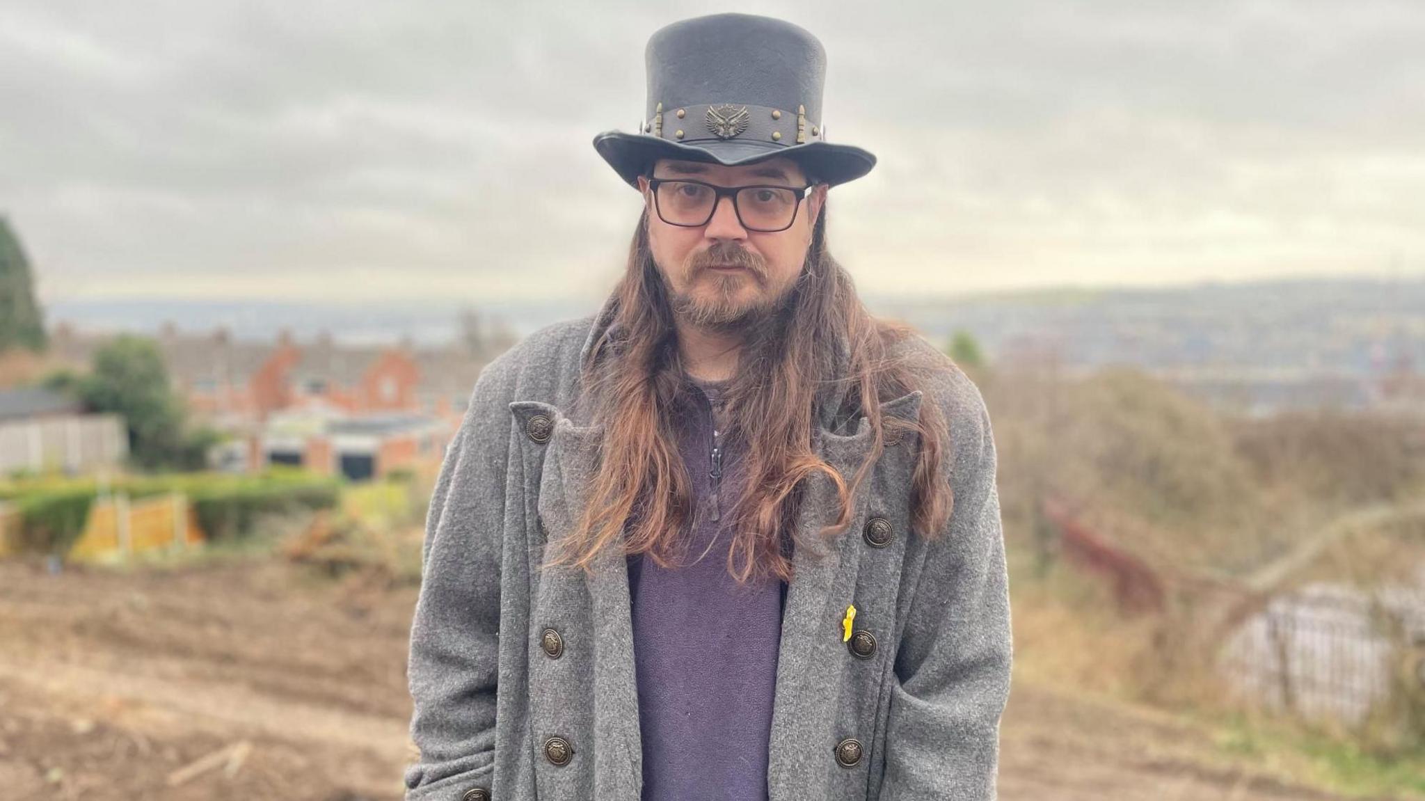 A man in a long grey coat and black hat stands with his back to an area of wasteland.