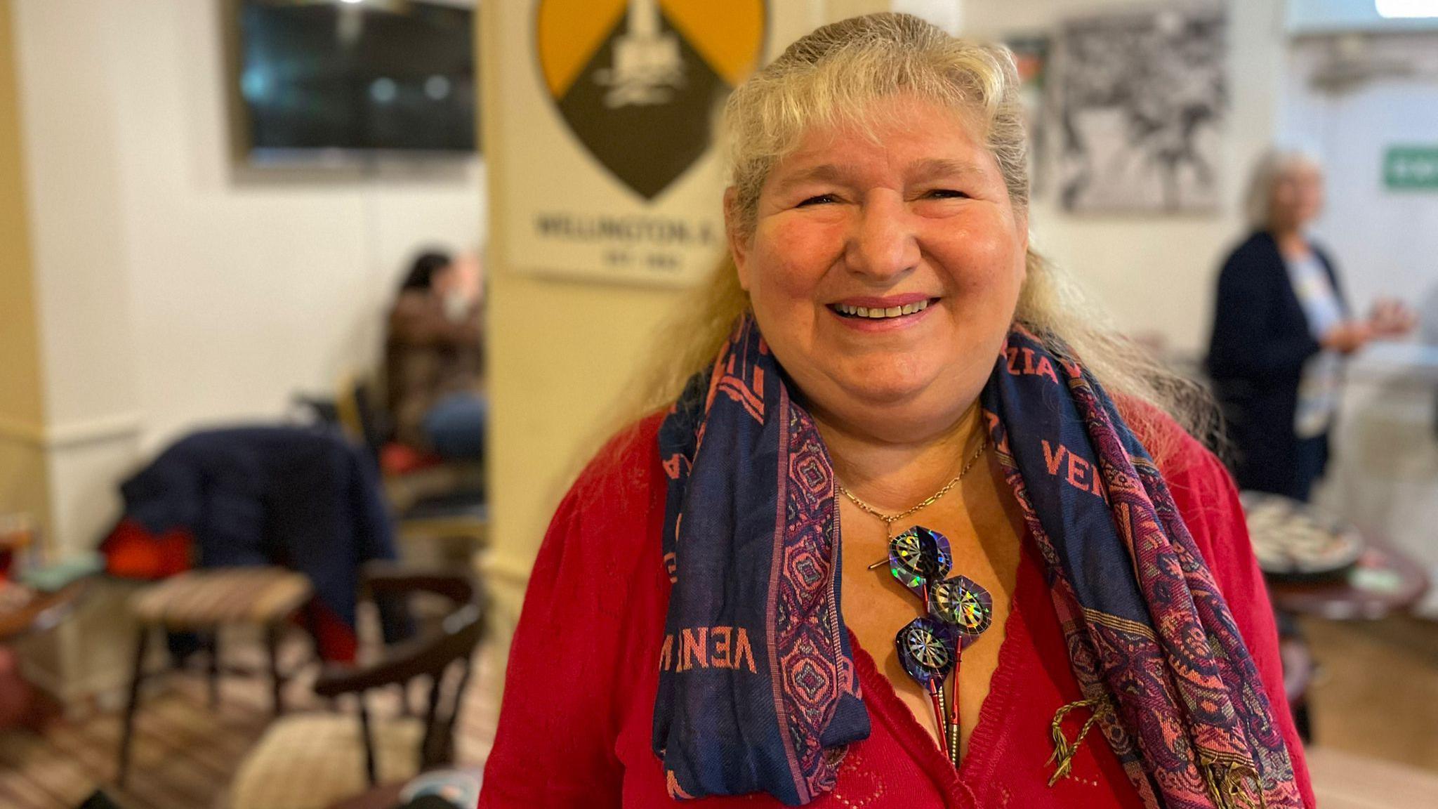 Ali is stood in the pub, smiling at the camera. She's wearing a red top with a blue scarf on. She has her darts poking out her top.