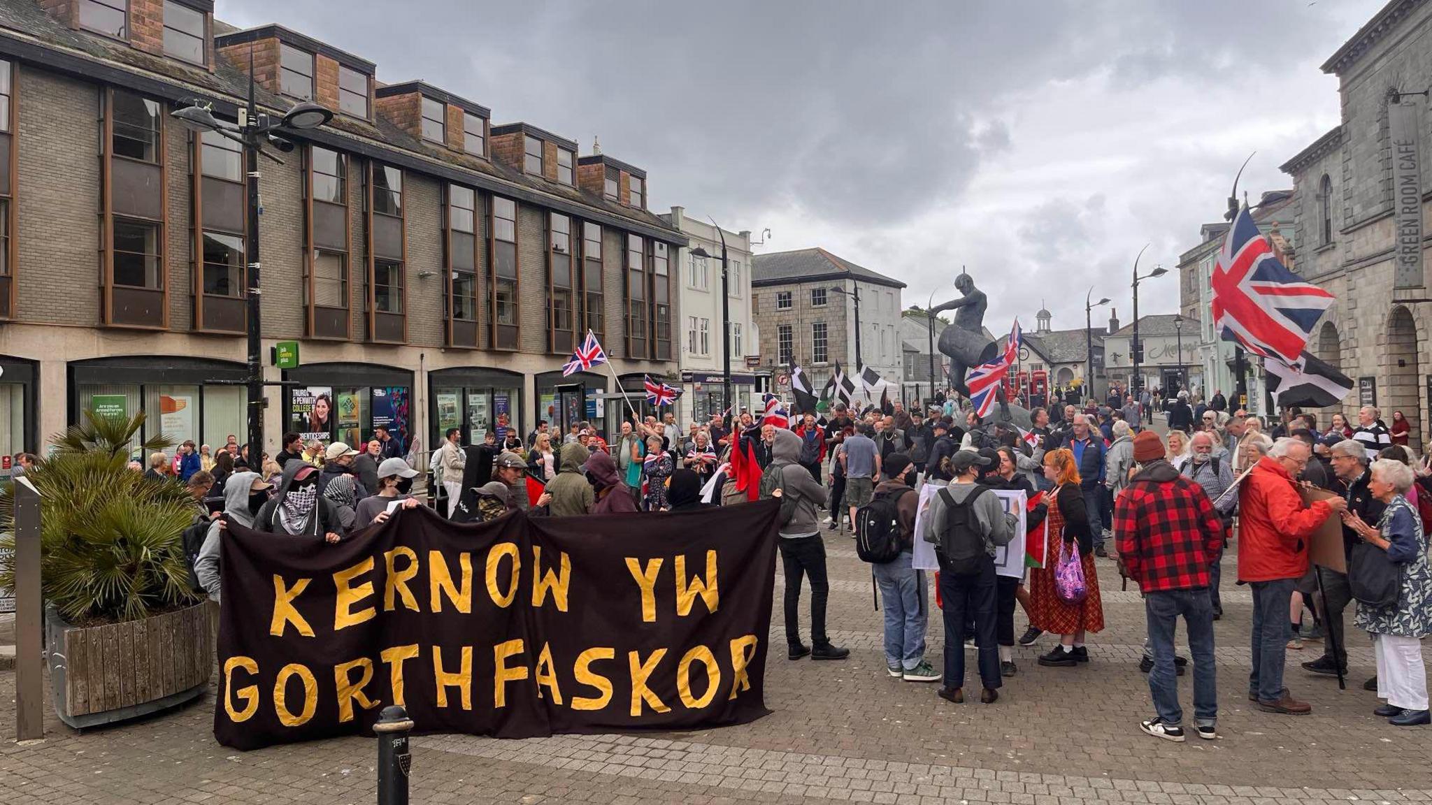 Crowds of people in Truro city centre with Union Jack flags and signs. There are buildings either side, and a statue towards the back. On the left, there are people holding a large black sign which has the words 'KERNOW YW GORTHFASKOR' in yellow writing.