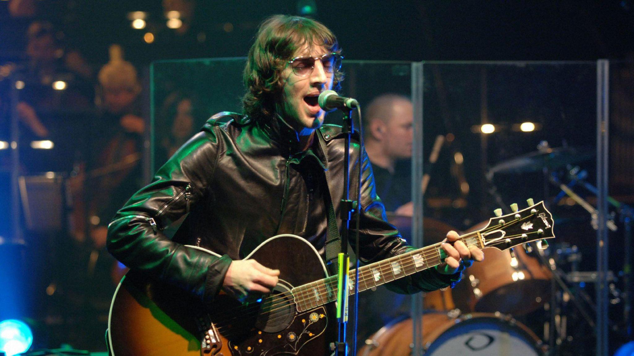 Richard Ashcroft performing with the BBC Concert Orchestra, holding a guitar and singing into a microphone, wearing sunglasses and a black leather jacket