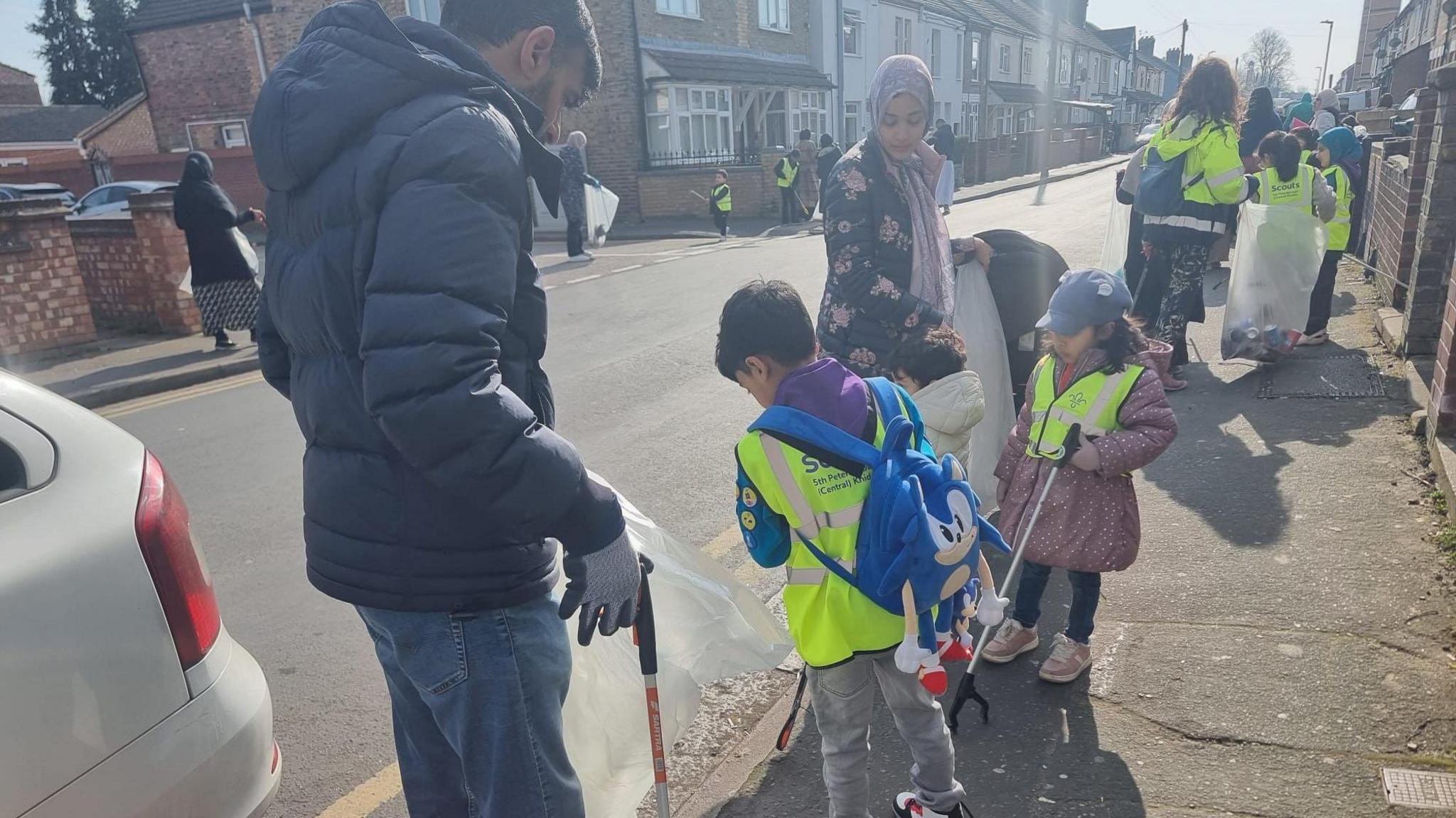 A man wearing a blue jacket and jeans holding a litter picker, with small children litter picking alongside a woman in a head scarf. In the background are other volunteers and terraced housing.