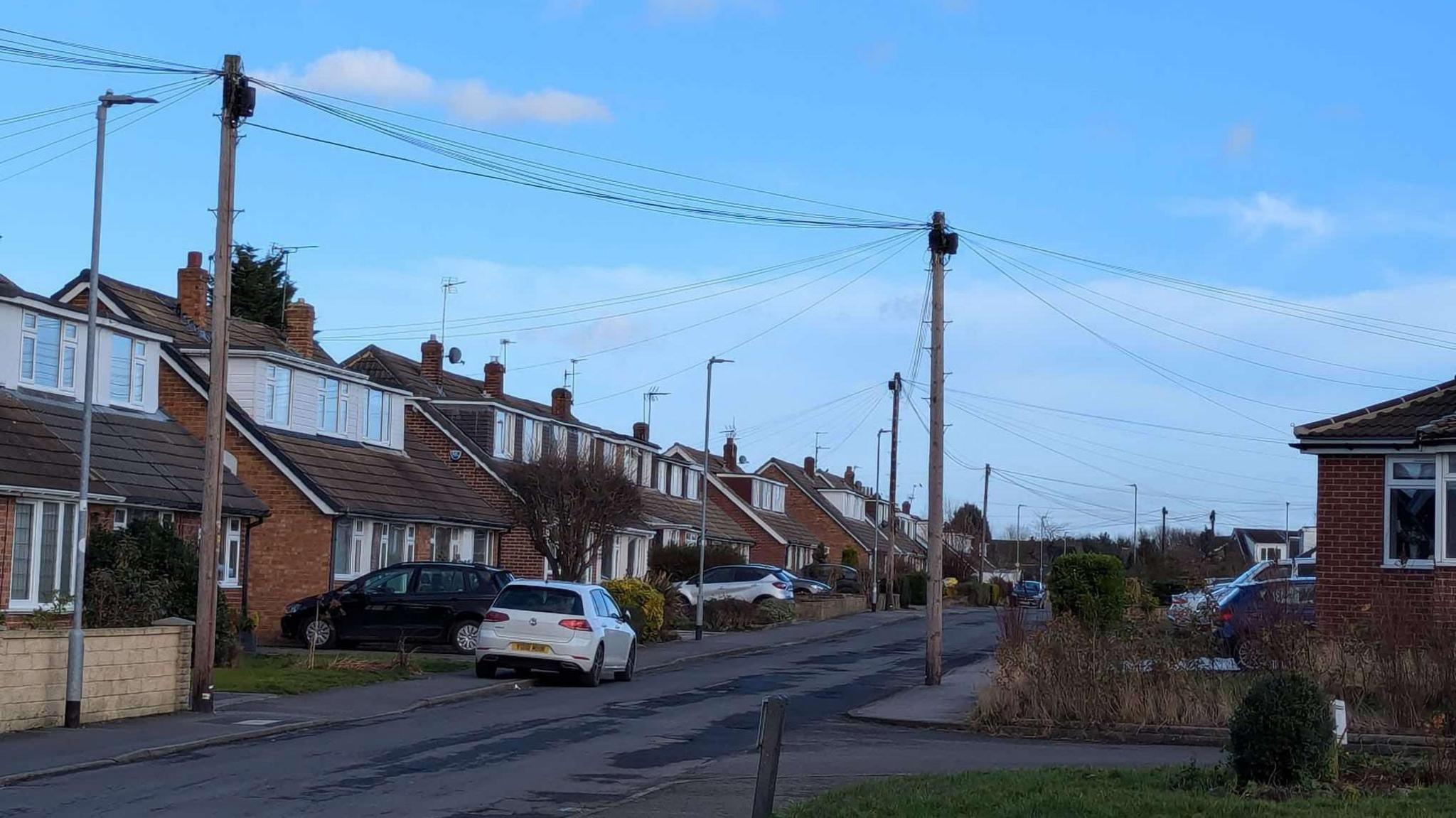 A street with houses and cars parked on the road and on driveways, showing telegraph poles with wires coming off them.