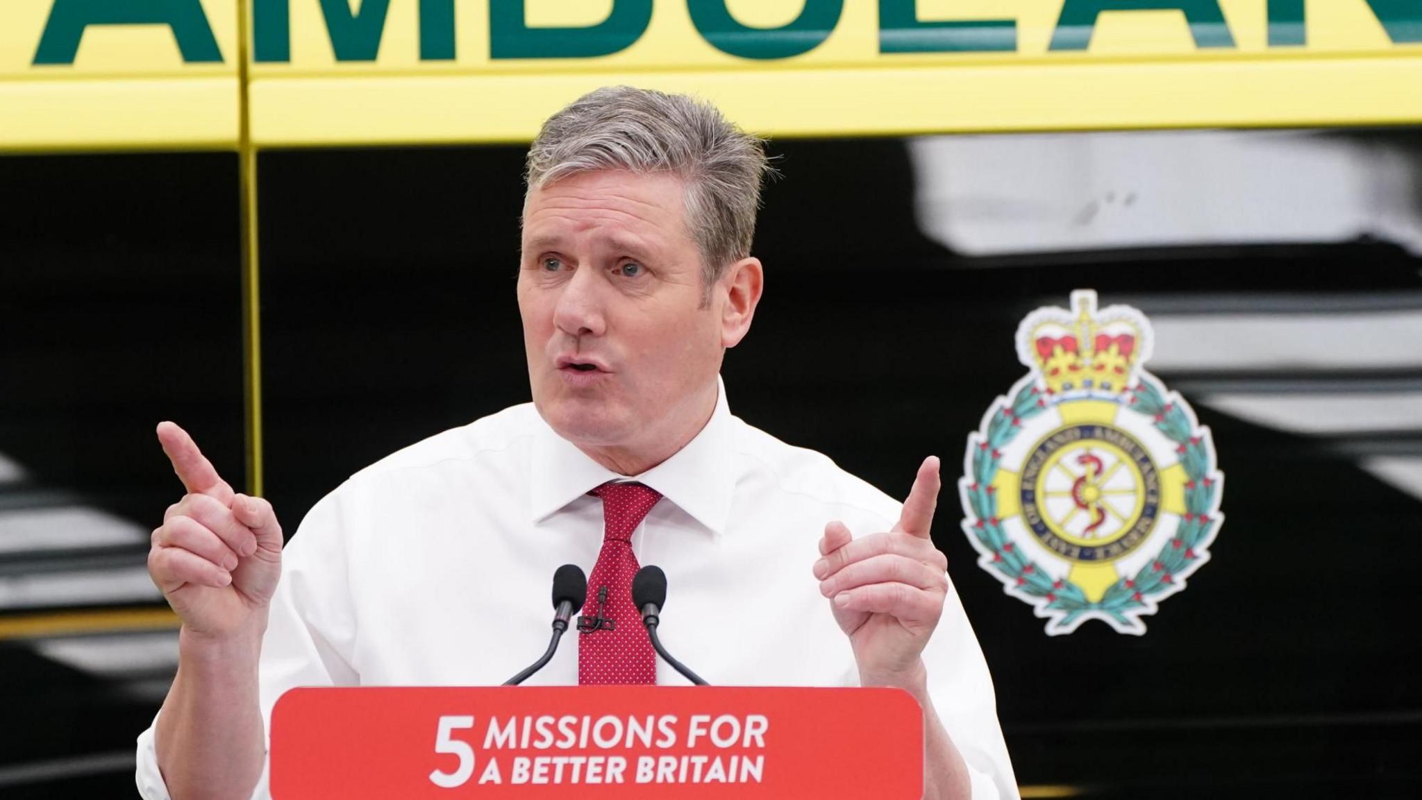 Sir Kier Starmer making a health speech at the East of England Ambulance Service Trust in Braintree in 2023. The picture shows Sir Keir holding up two pointed hands. He is wearing a white shirt and red tie. There is an ambulance in the background with a logo on it.  The lectern in front of him has the slogan "5 missions for a better britain".