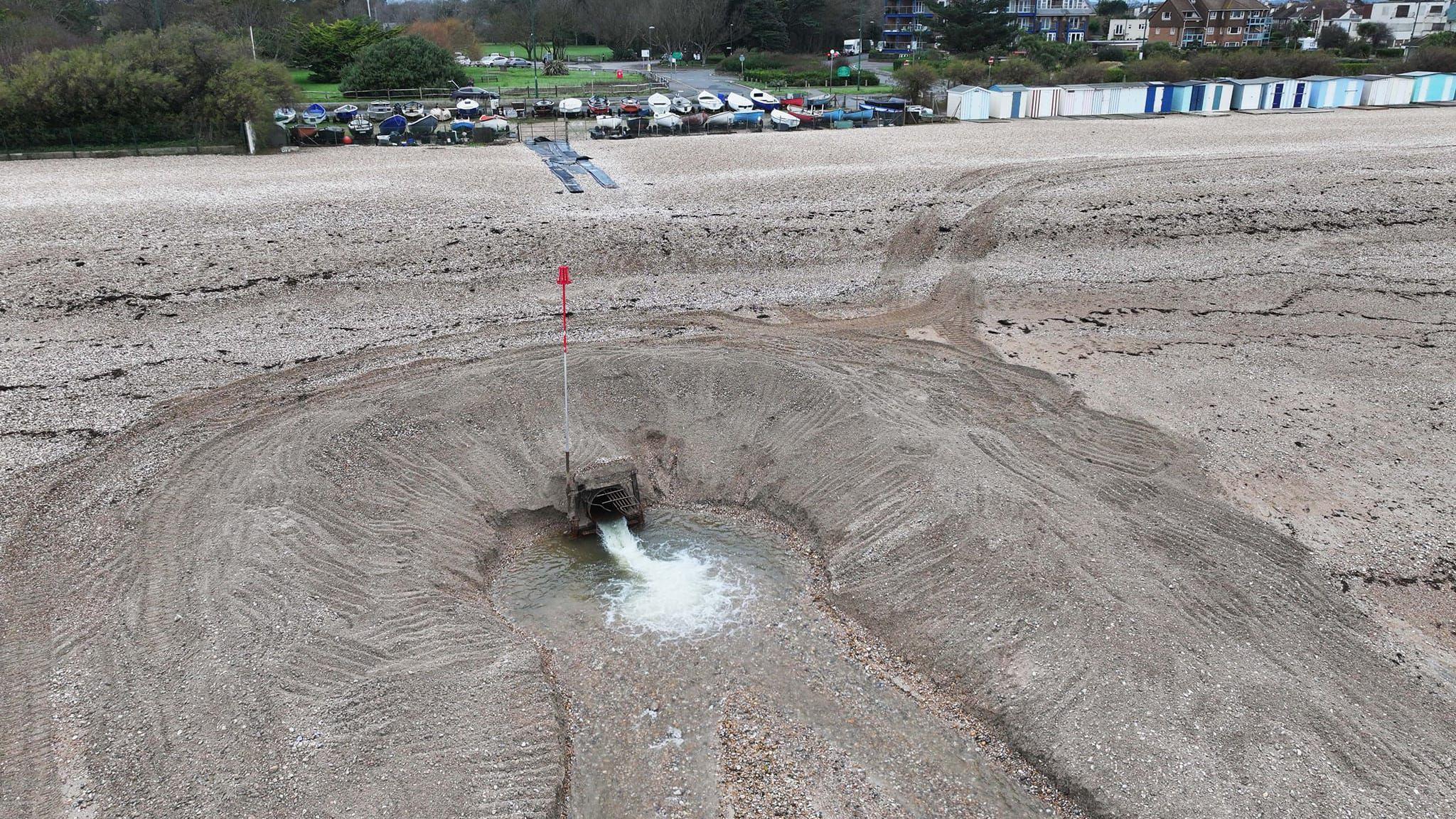 An aerial photo of an outfall pipe spewing out water with shingle around it about 100 metres away from Fish Lane which had flooding. The pipe is surrounded by a mound of shine and the water can be seen draining in two channels out to sea. Cars can be seen parked on the seafront and there are beach huts