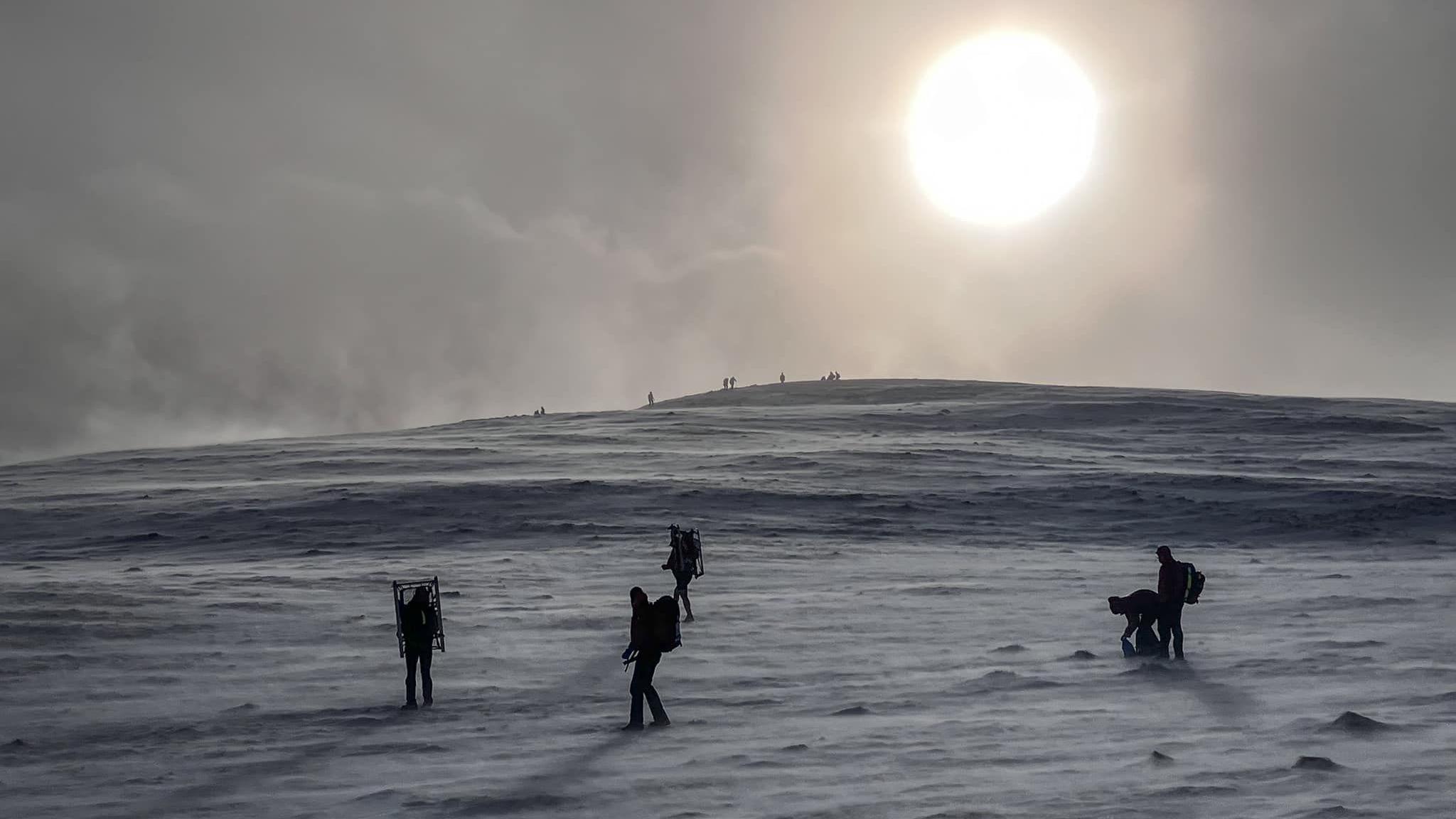 A bright sun shines a weak light over Blencathra which is covered in snow. There are five people silhouetted, wearing back packs and one carrying a stretcher. They are casting gentle shadows on the snow, which is almost grey-blue in the low light.  