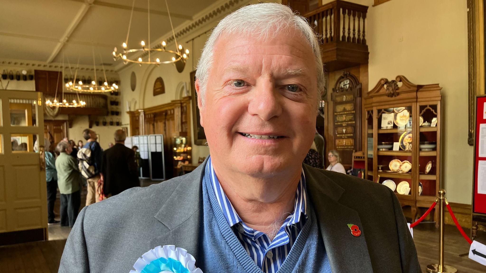 A man with short white hair smiles at the camera. He is wearing a blue and white striped shirt under a blue V-neck sweatshirt and a grey suit jacket. A poppy badge is on his left lapel and a blue and white rosette is on the other.