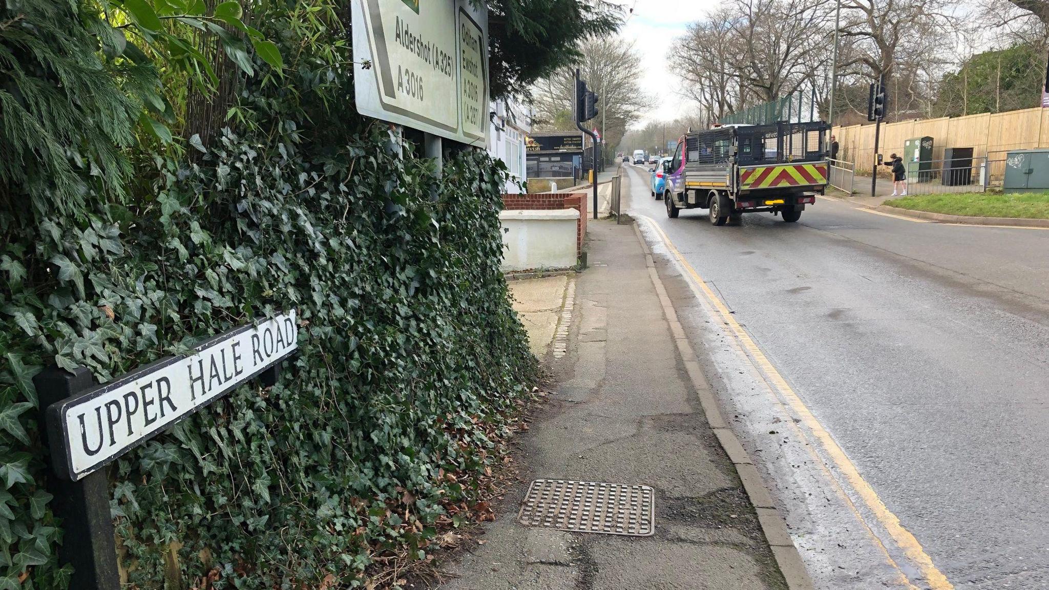 In the foreground a sign which says Upper Hale Road. Above it is some road direction signs. There is a pavement going alongside the road. There is also a large vehicle (although not an HGV) in the background.