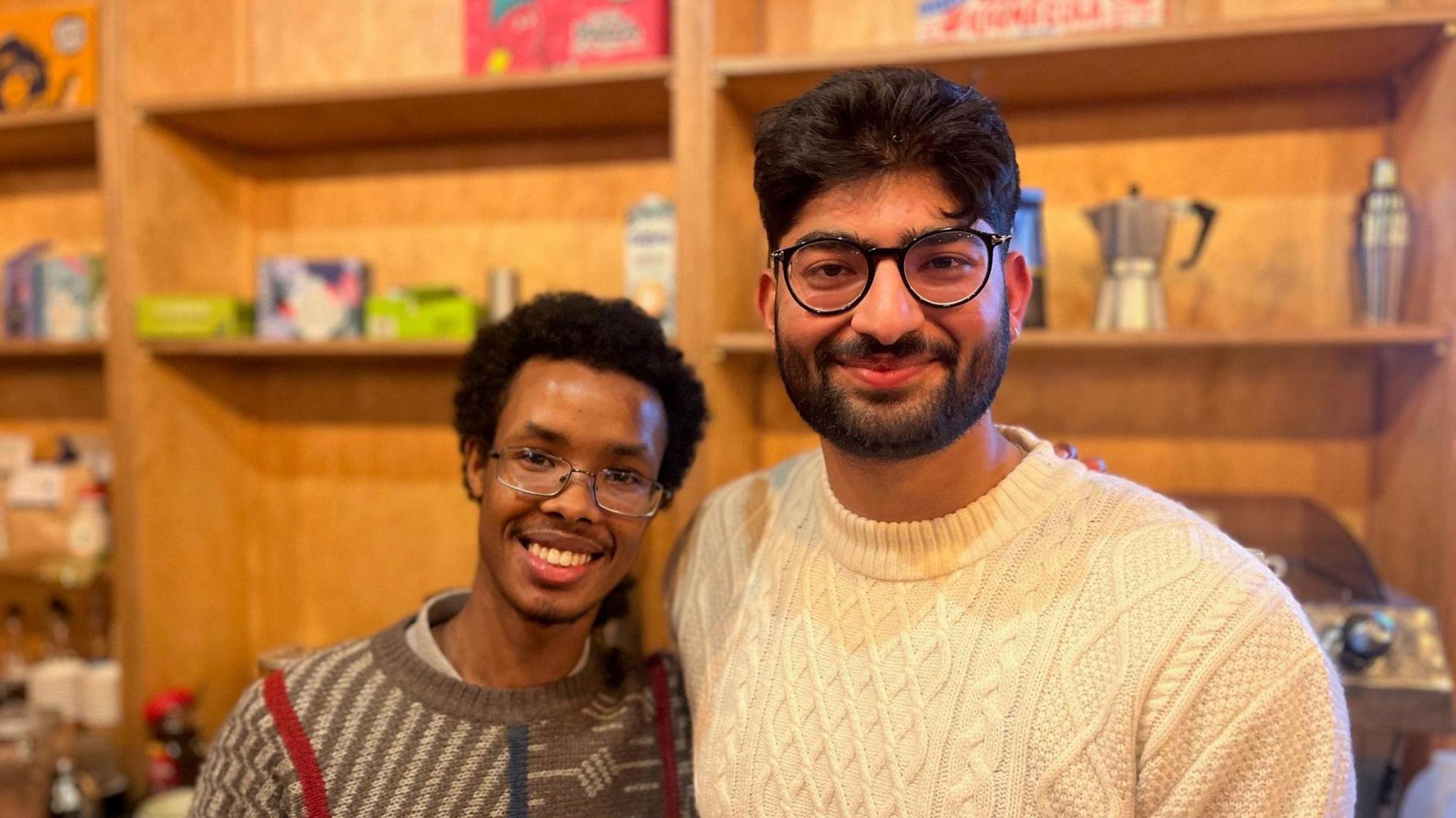 Two young men smile at the camera. Both have glasses, one is wearing a brown stripey jumper the other a cream one. Behing them are shelves filled with an assortment of products.