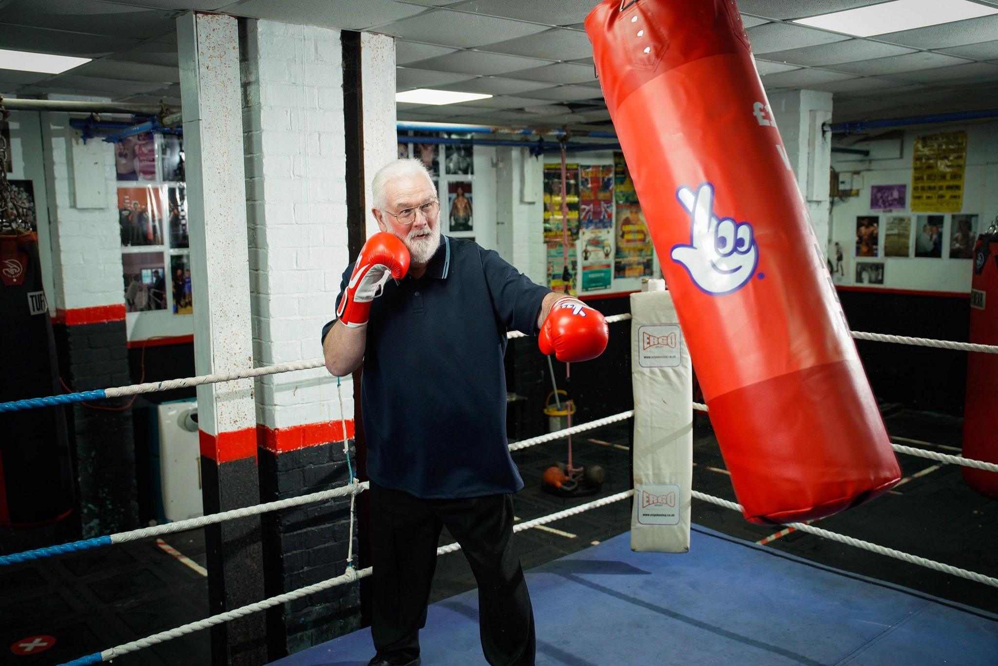 Peter Rogers stands in a boxing ring with red boxing gloves on hitting a red punching bag with the National Lottery logo. He has short white hair and short white beard. He is wearing glasses and a blue polo top. 