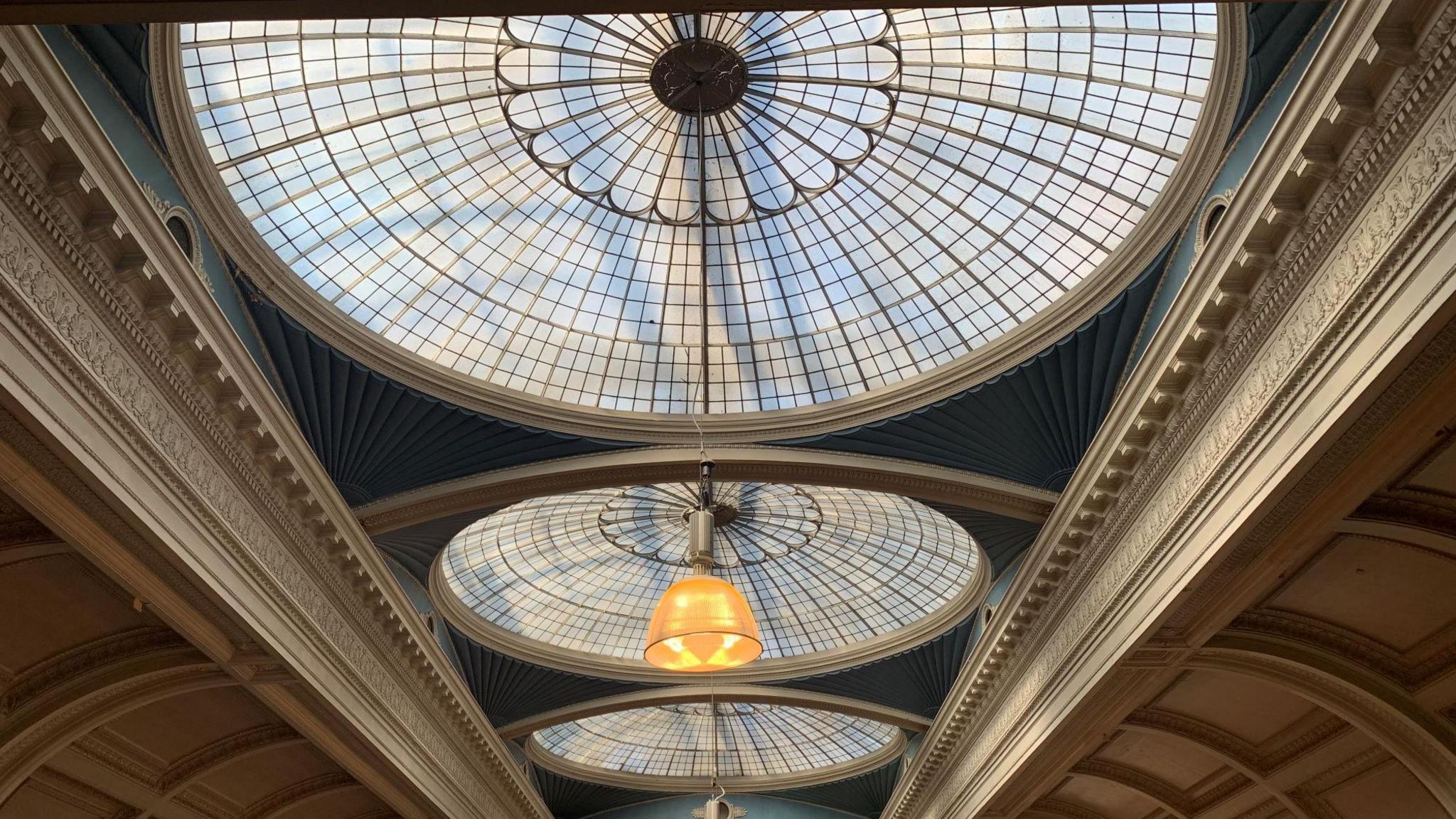 Three domed windows in a ceiling. The centre has a pendant lamp hanging down. To either side of the windows there is a white decorative panel on a ceiling.