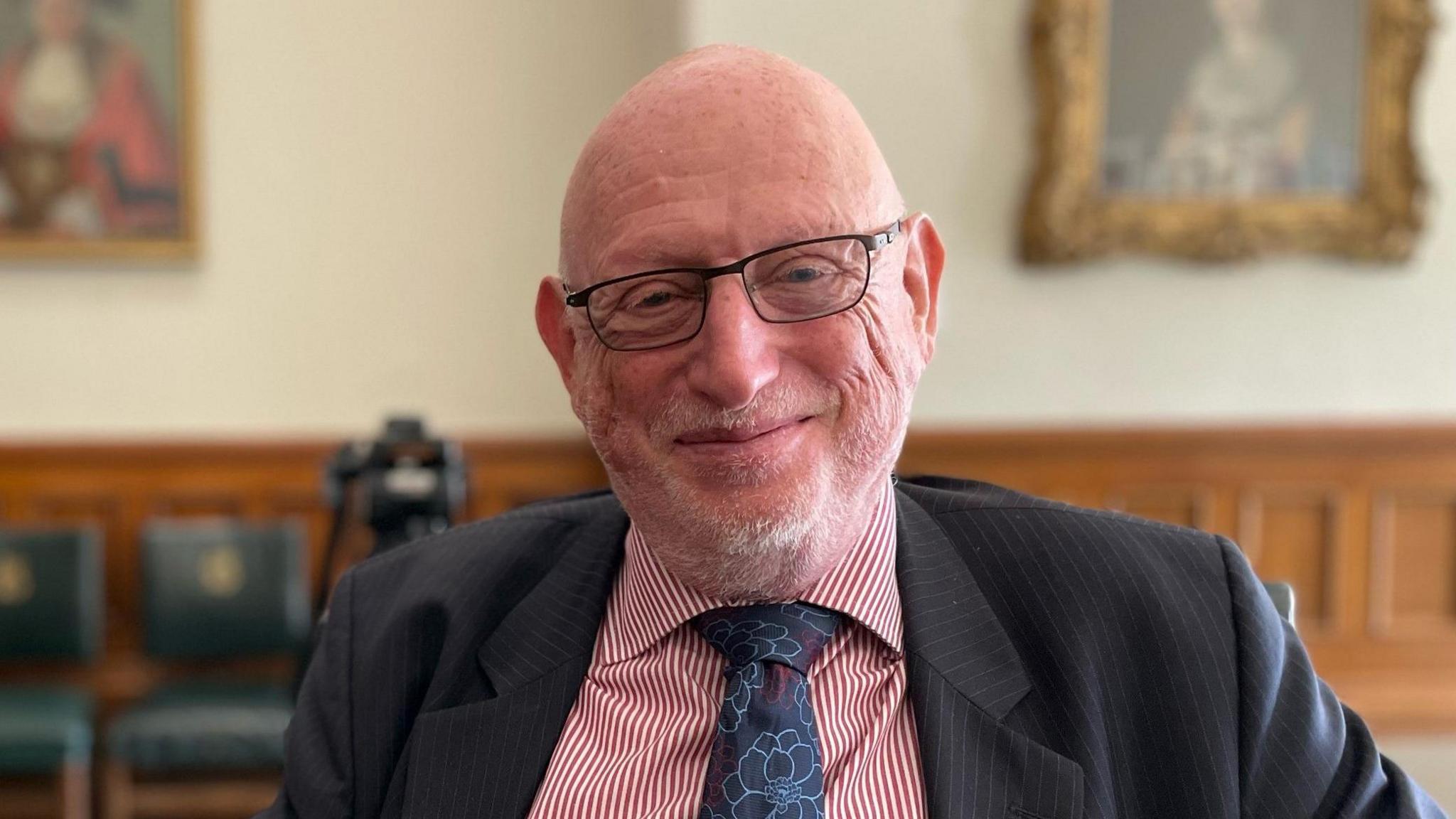 Max Caller wears glasses and a red and white pinstripe shirt. He has a dark blue tie with a floral pattern. He is wearing a dark grey pinstripe suit and is sat in an ornate room with wooden paneling.