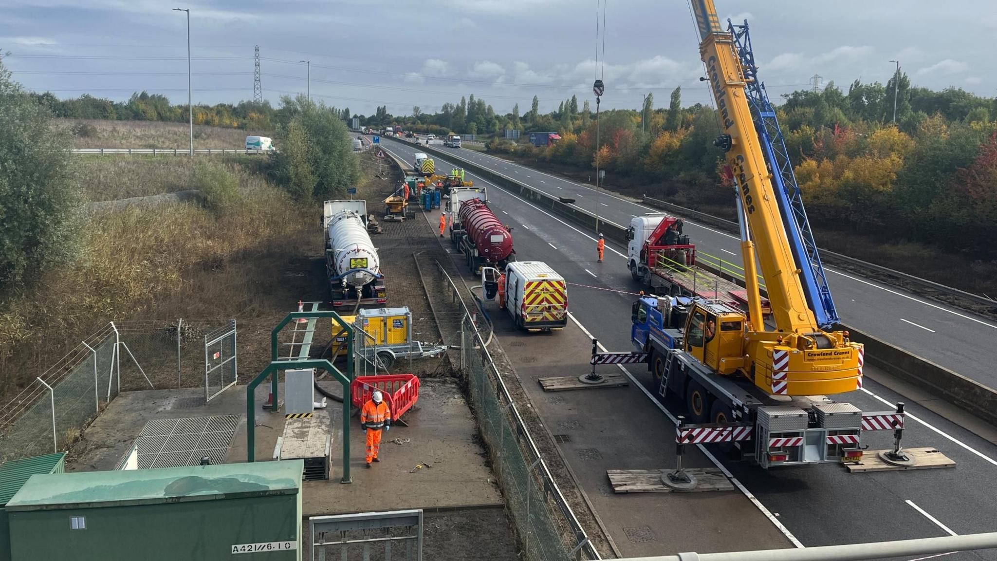 Men in orange high-vis jump suits work at the pumping station, a large crane is set up nearby. There are tankers, trucks and vans packed on the closed, but dry, A421.