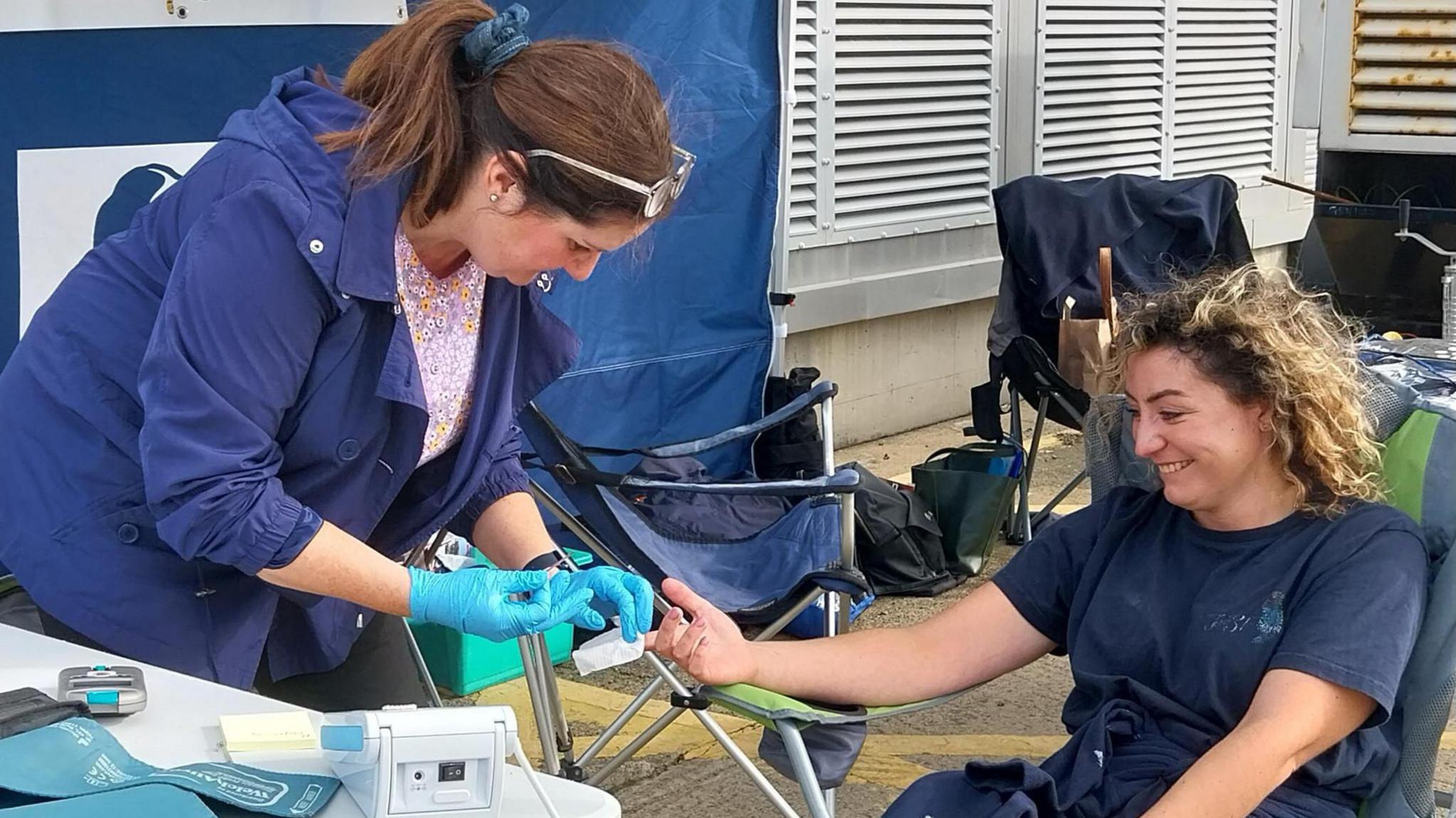 A woman is getting a health check from a physician. They are under a gazebo. The physician is wearing sterile gloves and holding a tissue over the other woman's finger- she's just had a blood test sample taken from her finger.