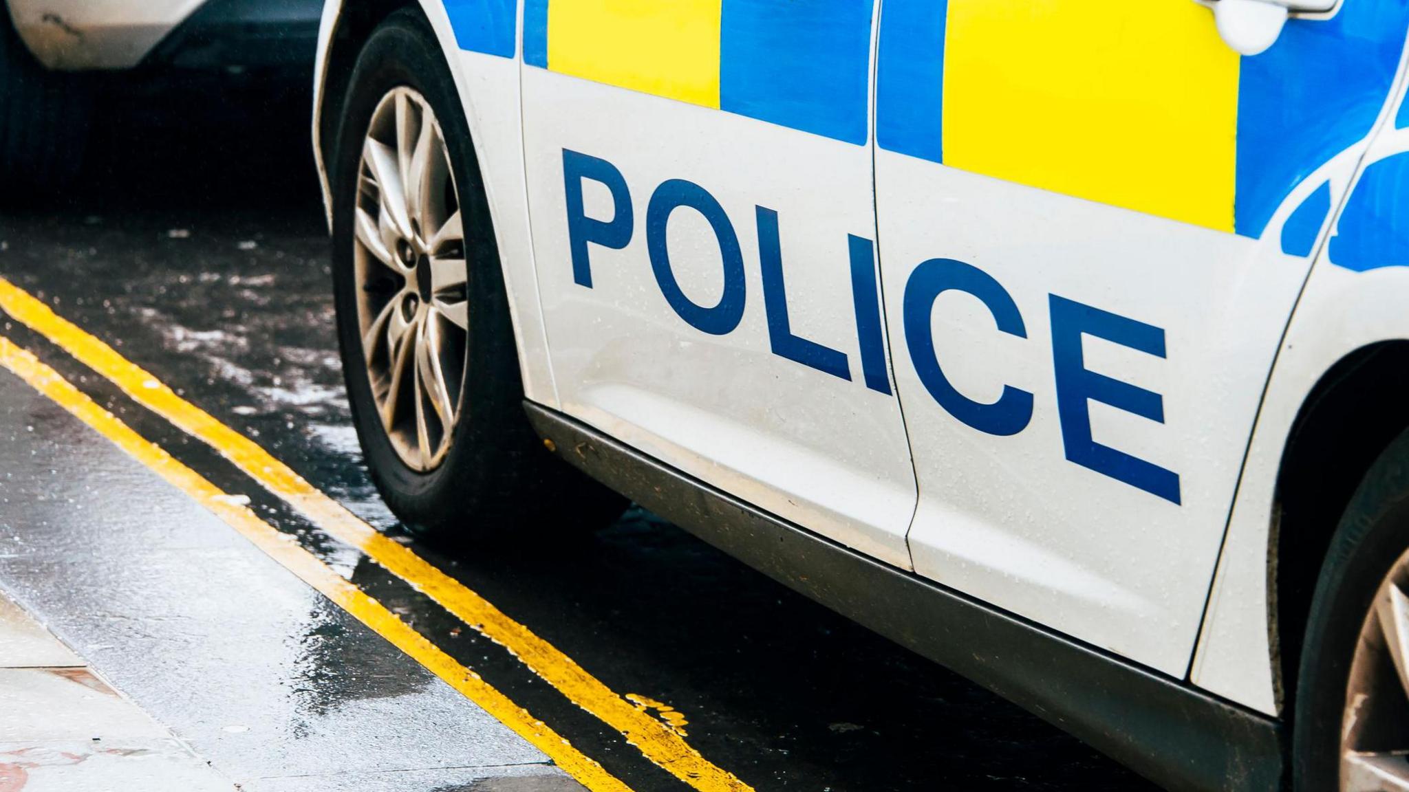 The detail of a police car. Dirty wheels can be seen on a wet pavement with double yellow lines. The car says police on the side. It has hi-vis markings. 