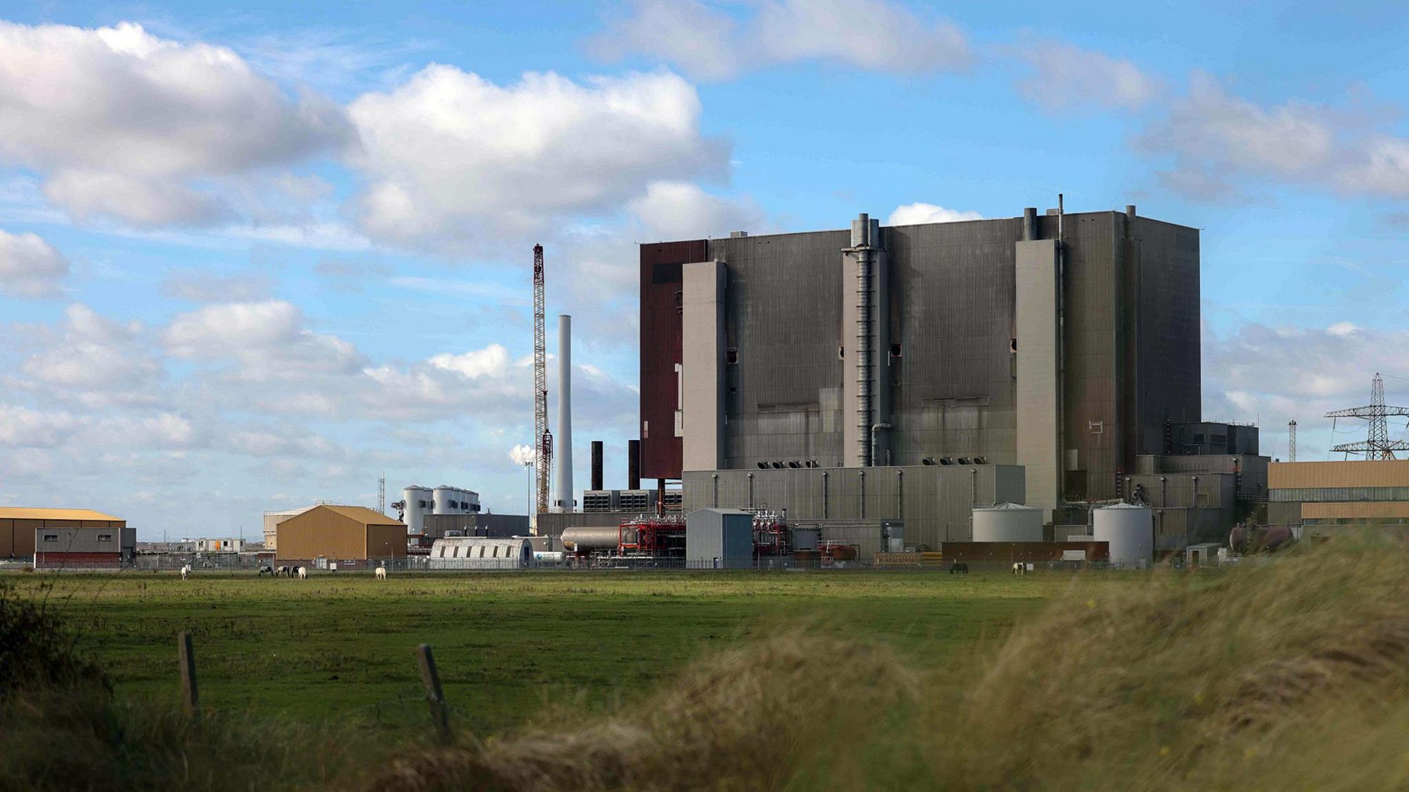 Hartlepool nuclear power station - a dark grey building surrounded by towers and pylons - situated on a stretch of grass with a fence and foliage in the foreground.