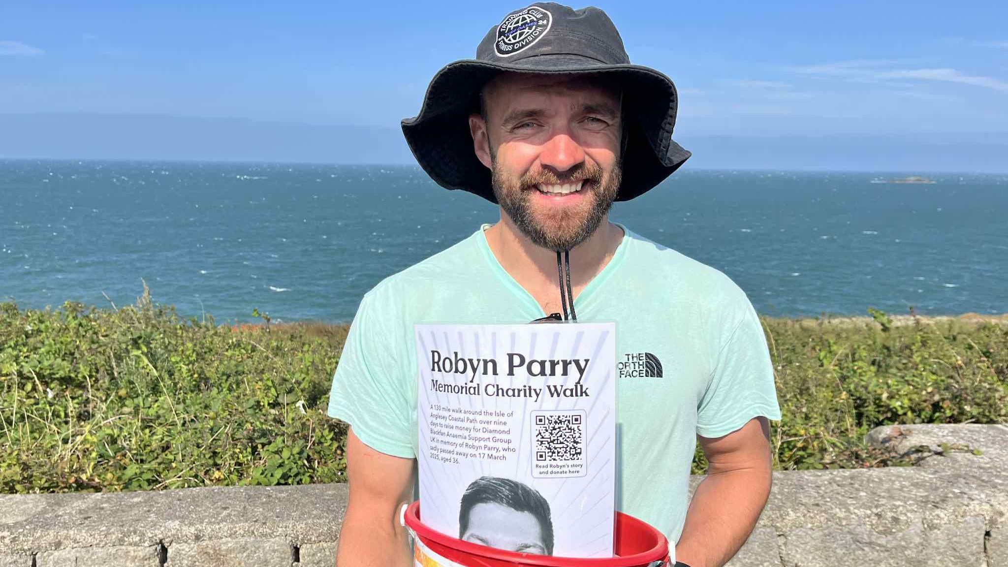 Ben Brame holding a red collection bucket with a piece of paper in it titled 'Robyn Parry memorial charity walk. He is wearing a grey bucket hat and a light green t-short, and has a short black beard. The sea can be seen behind him.