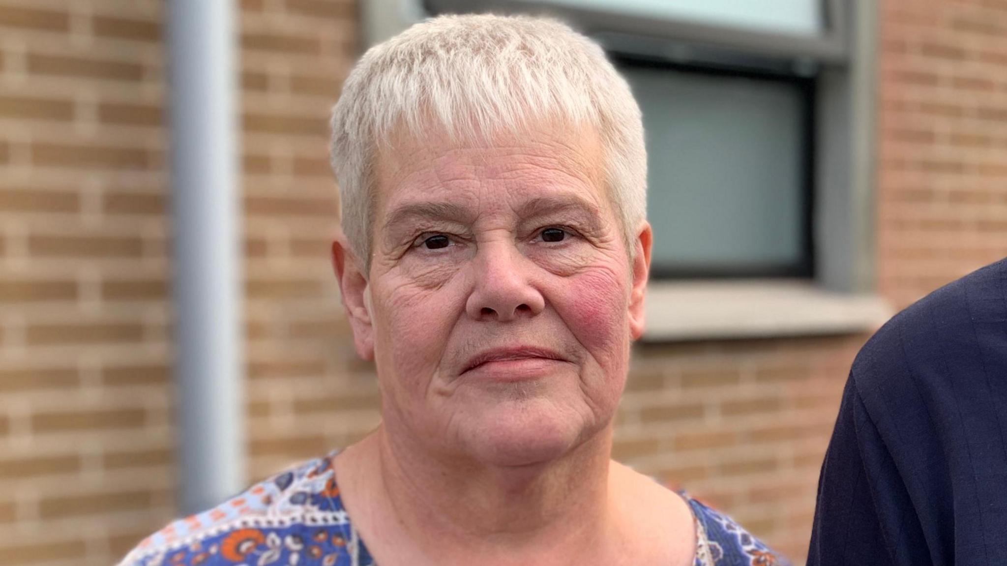A woman with short white hair and brown eyes looks into the camera. Cut off at the shoulder you can see she wears a blue, orange and white coloured top. A mans shoulder can be seen to her left, he wears a navy top. Behind her, out-of-focus is a red bricked wall. A partially opened translucent window is positioned over her left shoulder. 