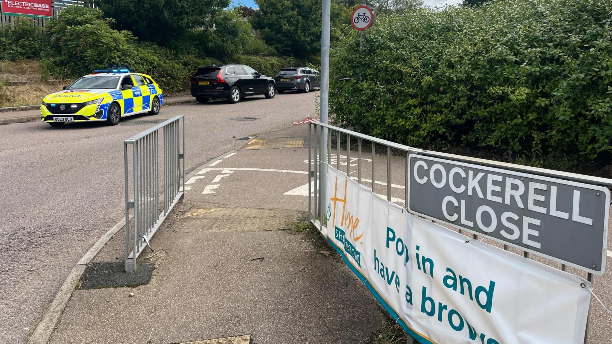 A road with a police car, black car and silver car parked alongside its left-hand pavement. On the other side of the road the pavement meets a cycle path, which has green bushes running alongside it. The pavement has silver railings and bears a sign reading "Cockerell Close".