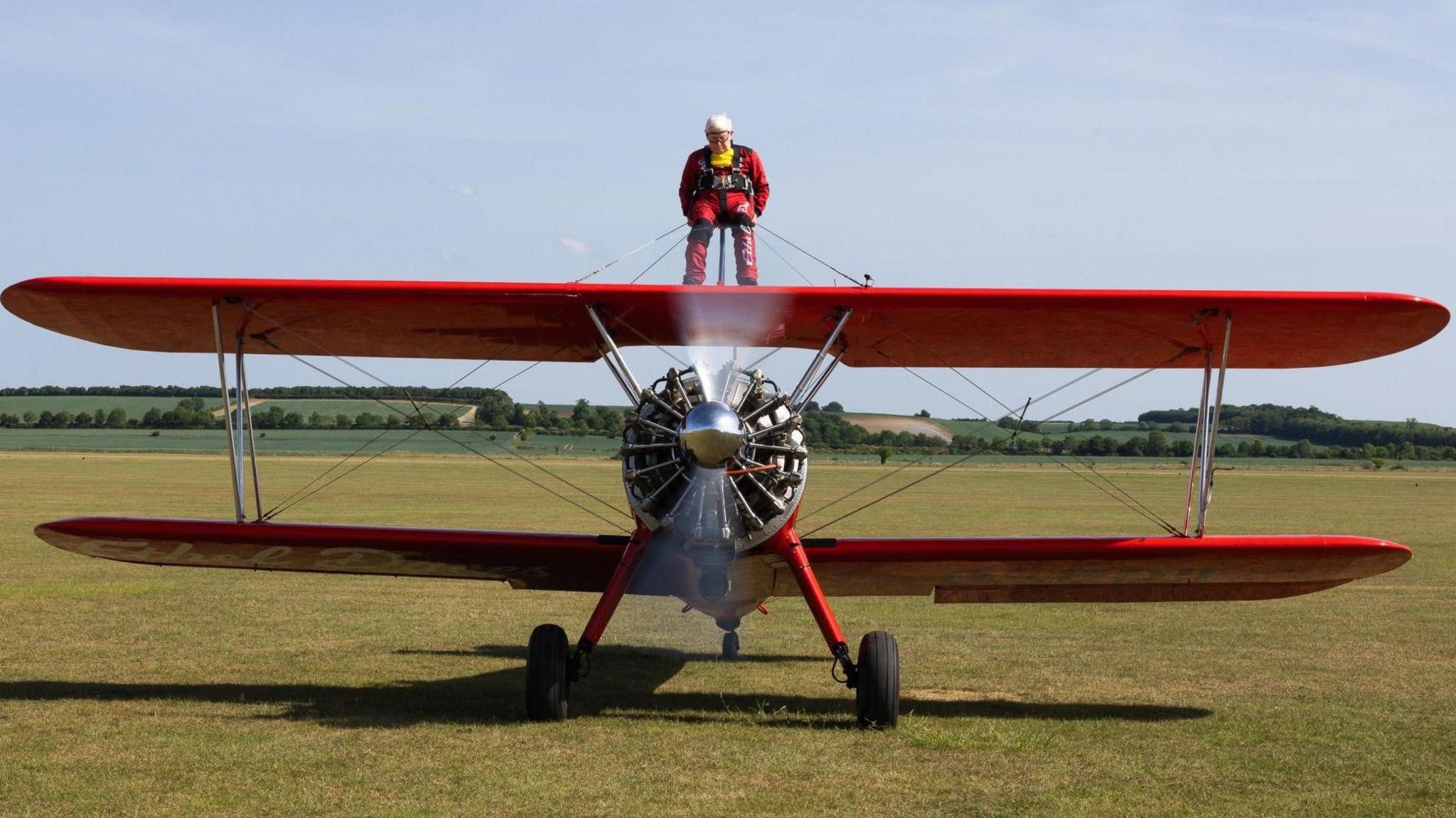Harry is wearing a red and black jump suit and is standing on top of a red plane, in the middle of a grass landing strip at the airfield.