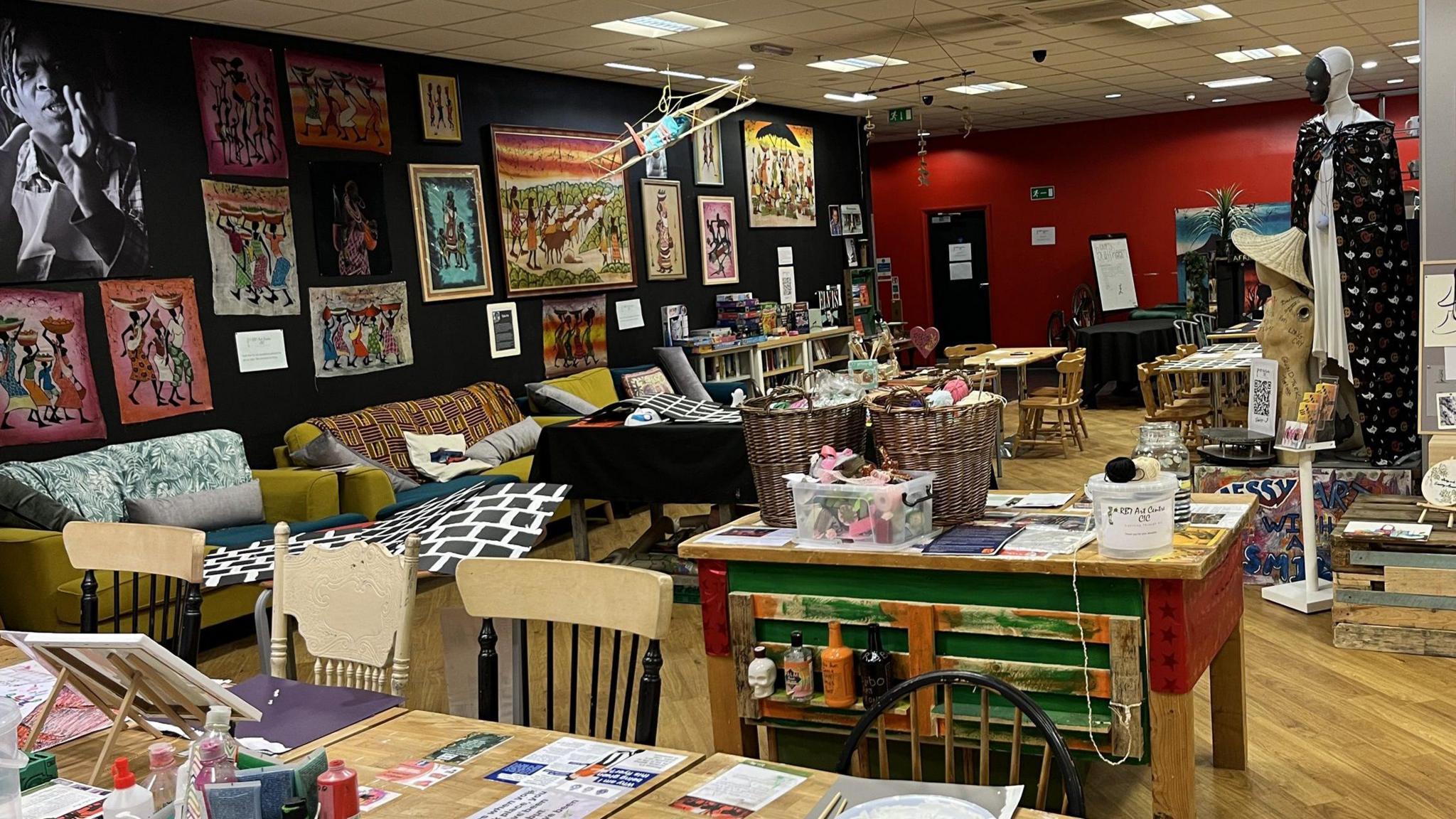 A room with chairs and tables laden with art supplies. There are also sofas and mannequins. There are several paintings hanging on the wall. The room appears busy and colourful.