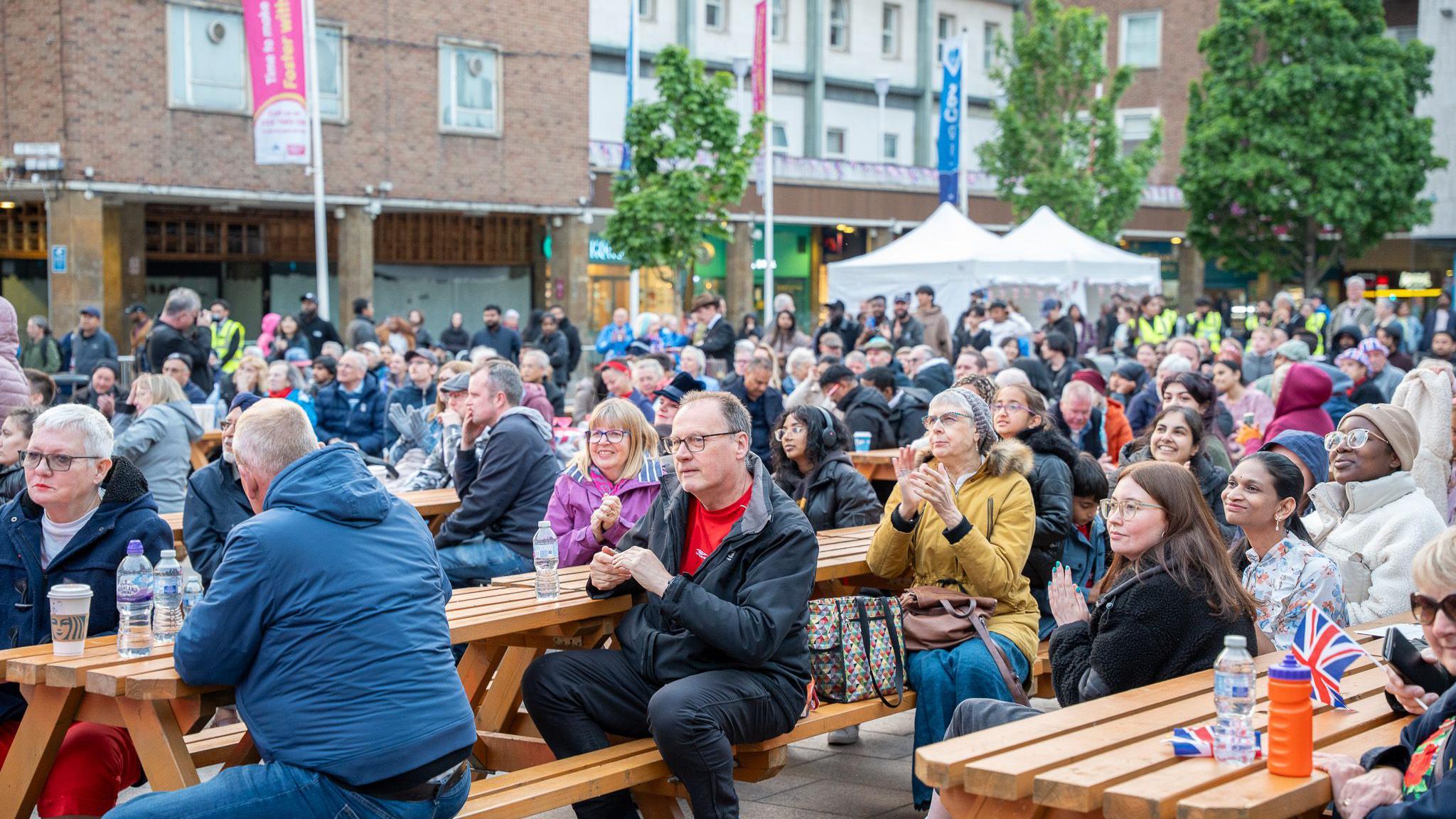 People sit at picnic tables, some holding Union Hack flags, and most wearing coats and with drinks like bottled water and coffee. There are two white tents in the background and buildings and trees in the distance.