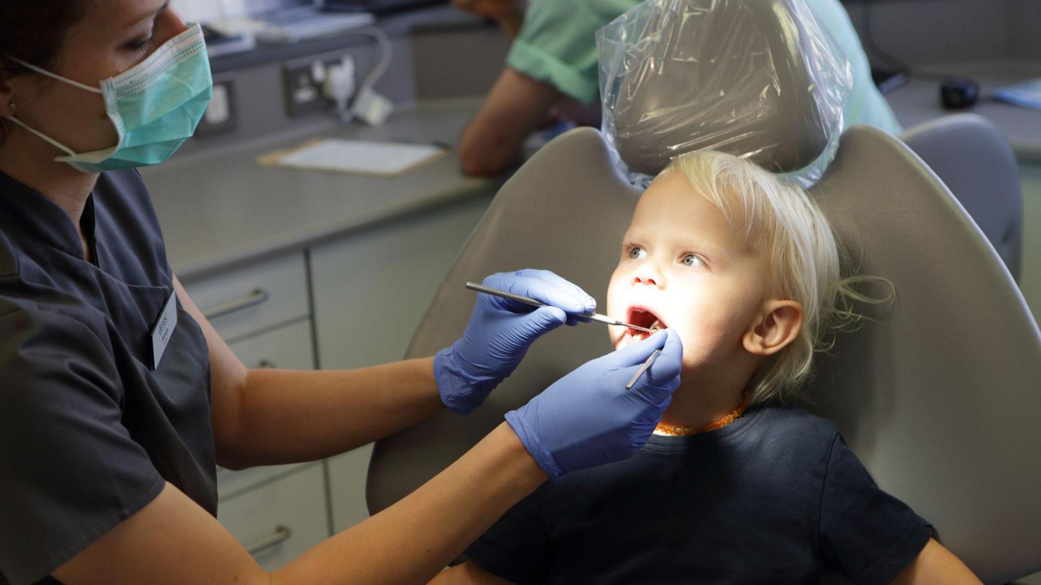 A young blonde boy sat in a dentist chair. He is looking at a female dentist who is looking in his mouth with two dental tools.