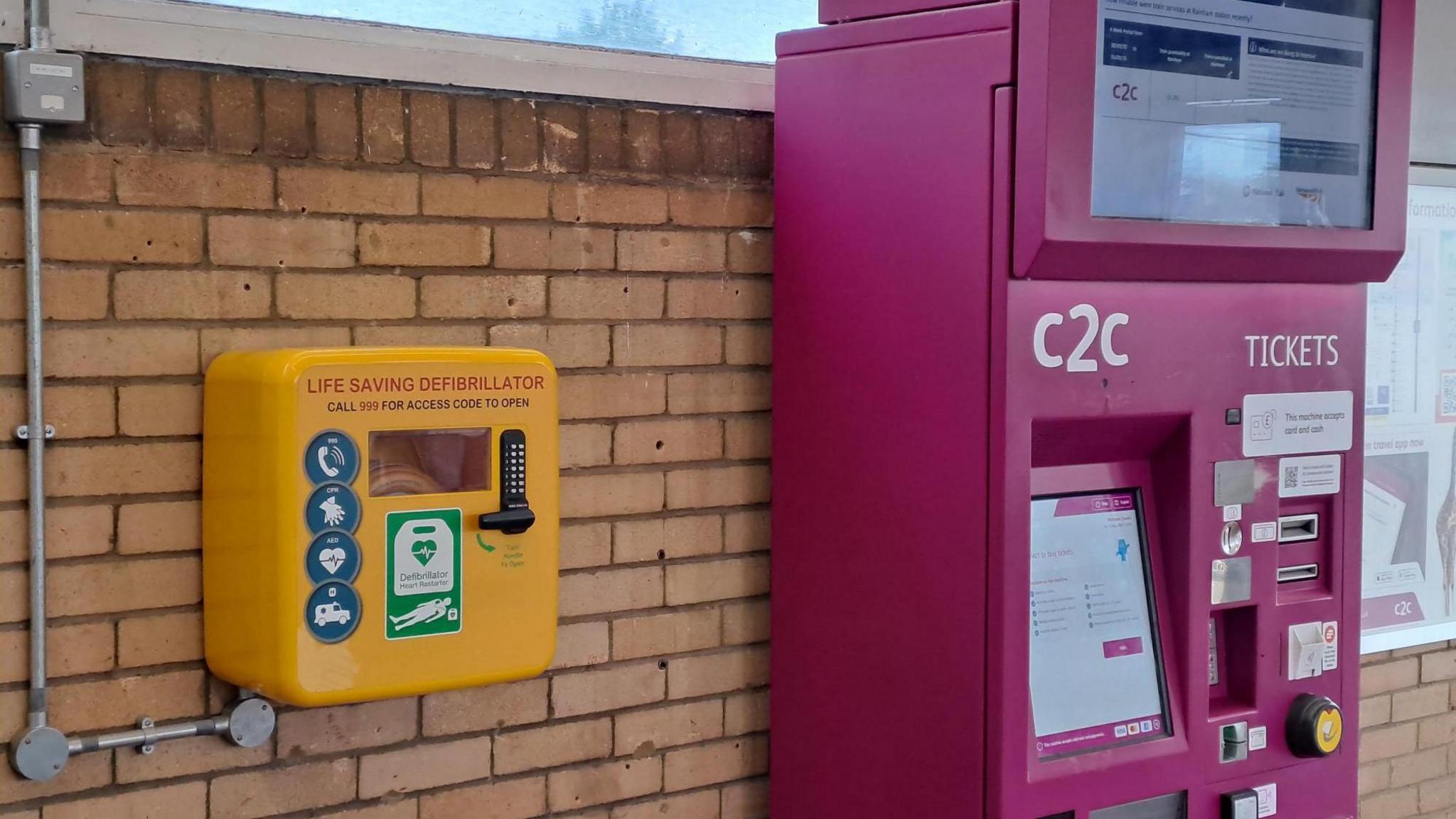A bright yellow defibrillator box and a larger purple ticket machine affixed to a brick wall