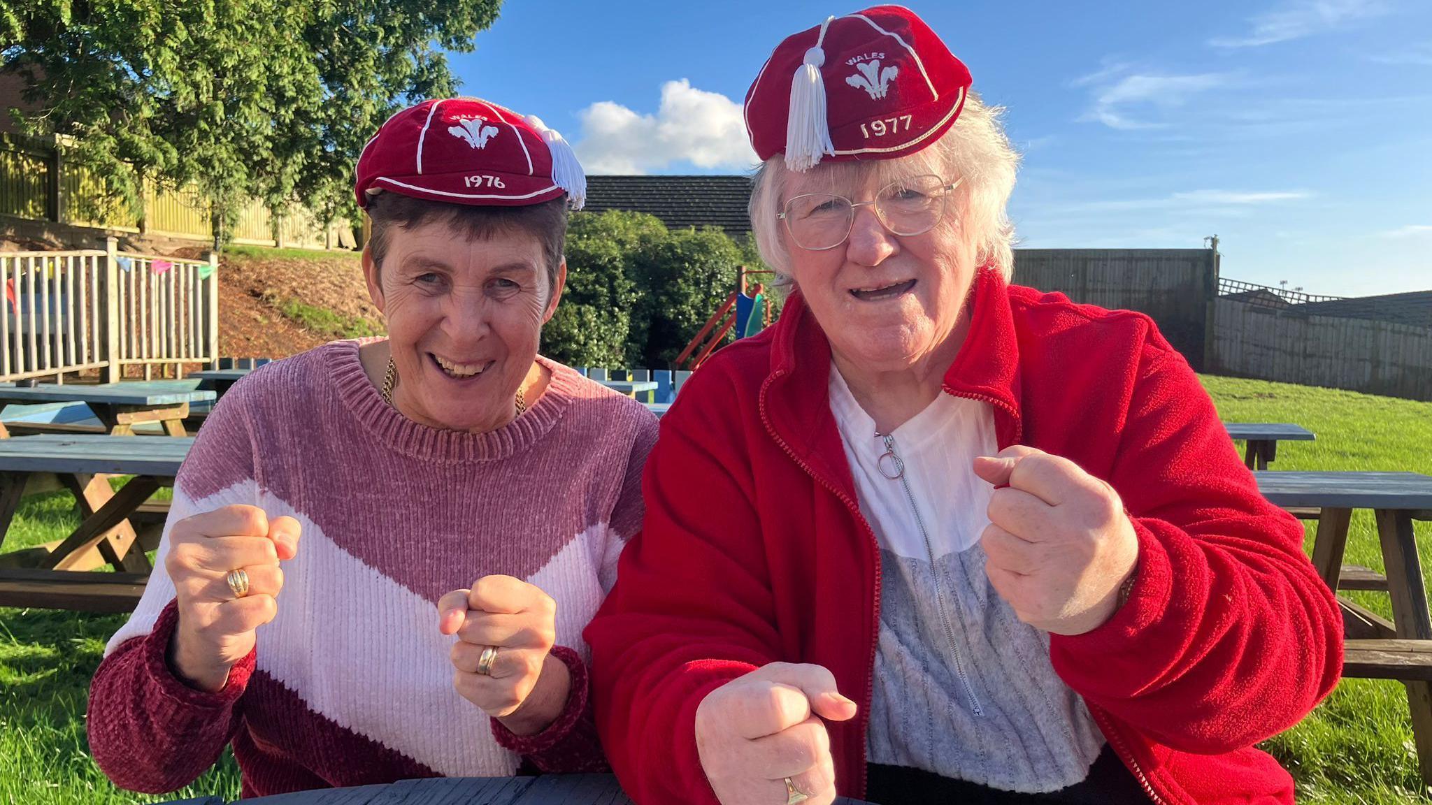 Gloria and Jayne sit at a picnic table clasping their fists in a show of celebration and hope, wearing their caps