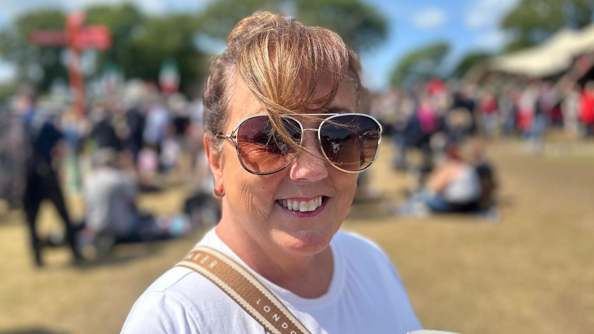 Ann Campbell wears a white t-shirt, and her hair in a bun. Her fringe is being blown over her sunglasses, she smiles.