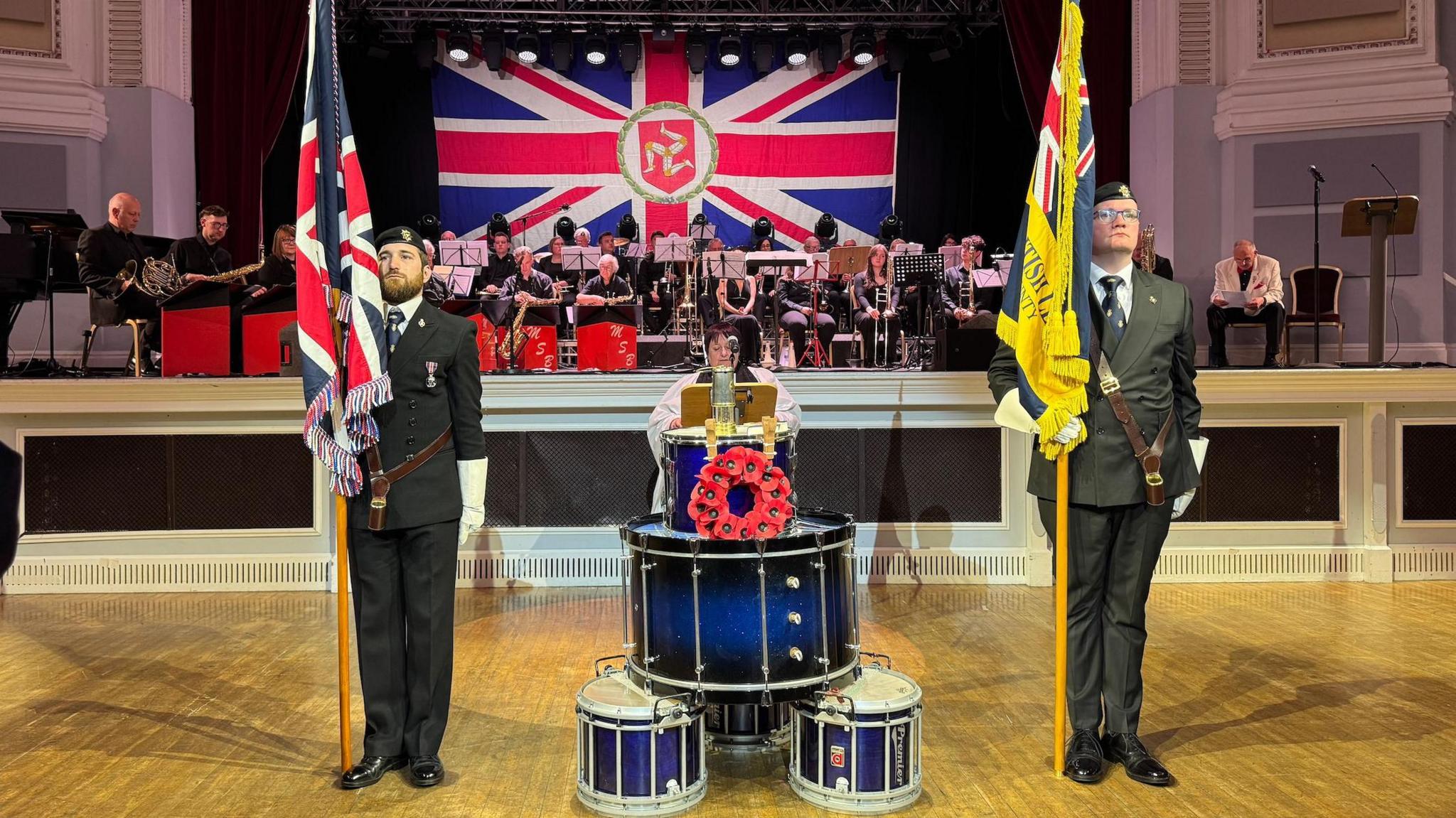 Two men standing in military uniforms and holding up flags n the Villa Marina Royal Hall. They are standing next to blue and white drums, which has a red wreath been placed on one, and there is a large band sitting behind them.