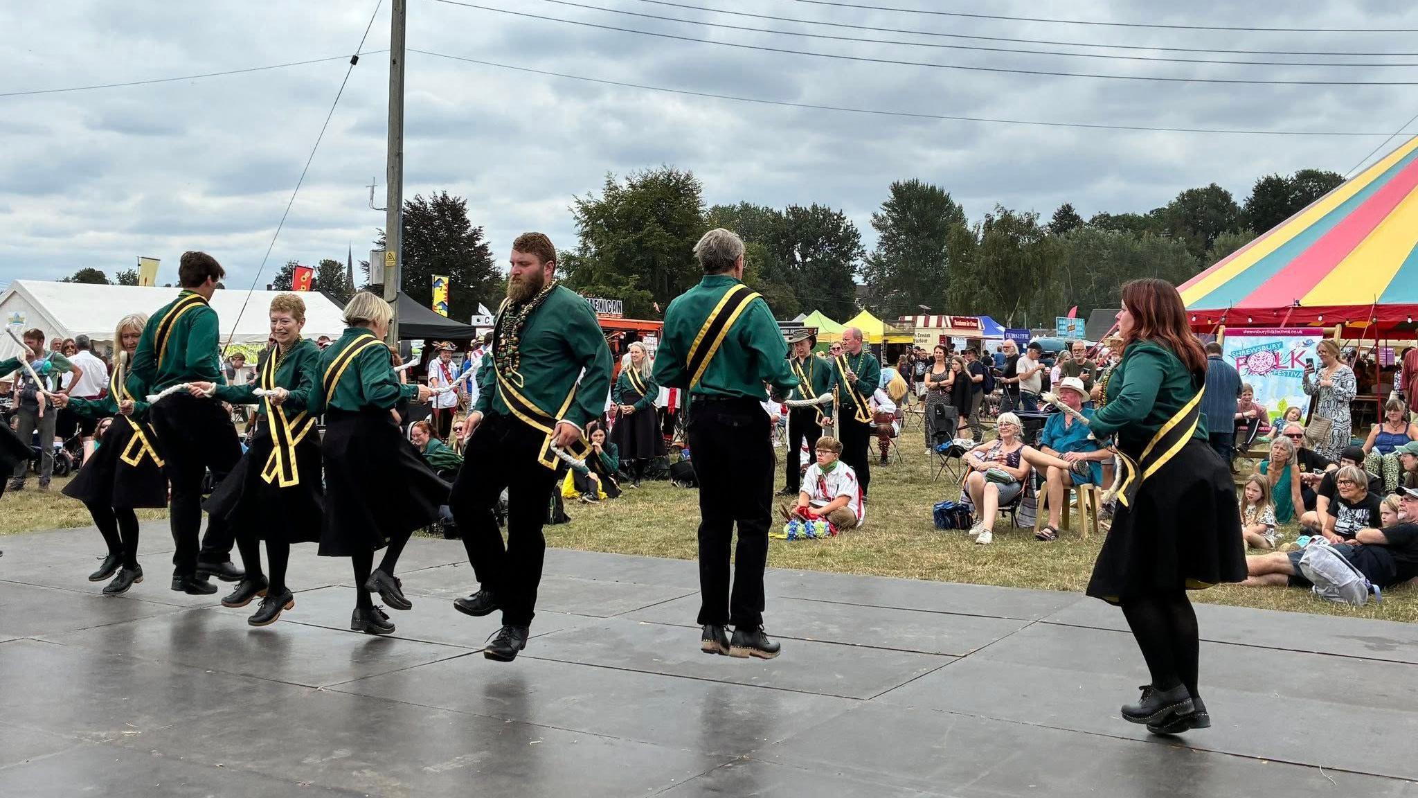 Morris dancers dressed in bottle green shirts and black trousers performing on a stage made out if black mats.  A crowd are gathered in front sitting on camping chairs.