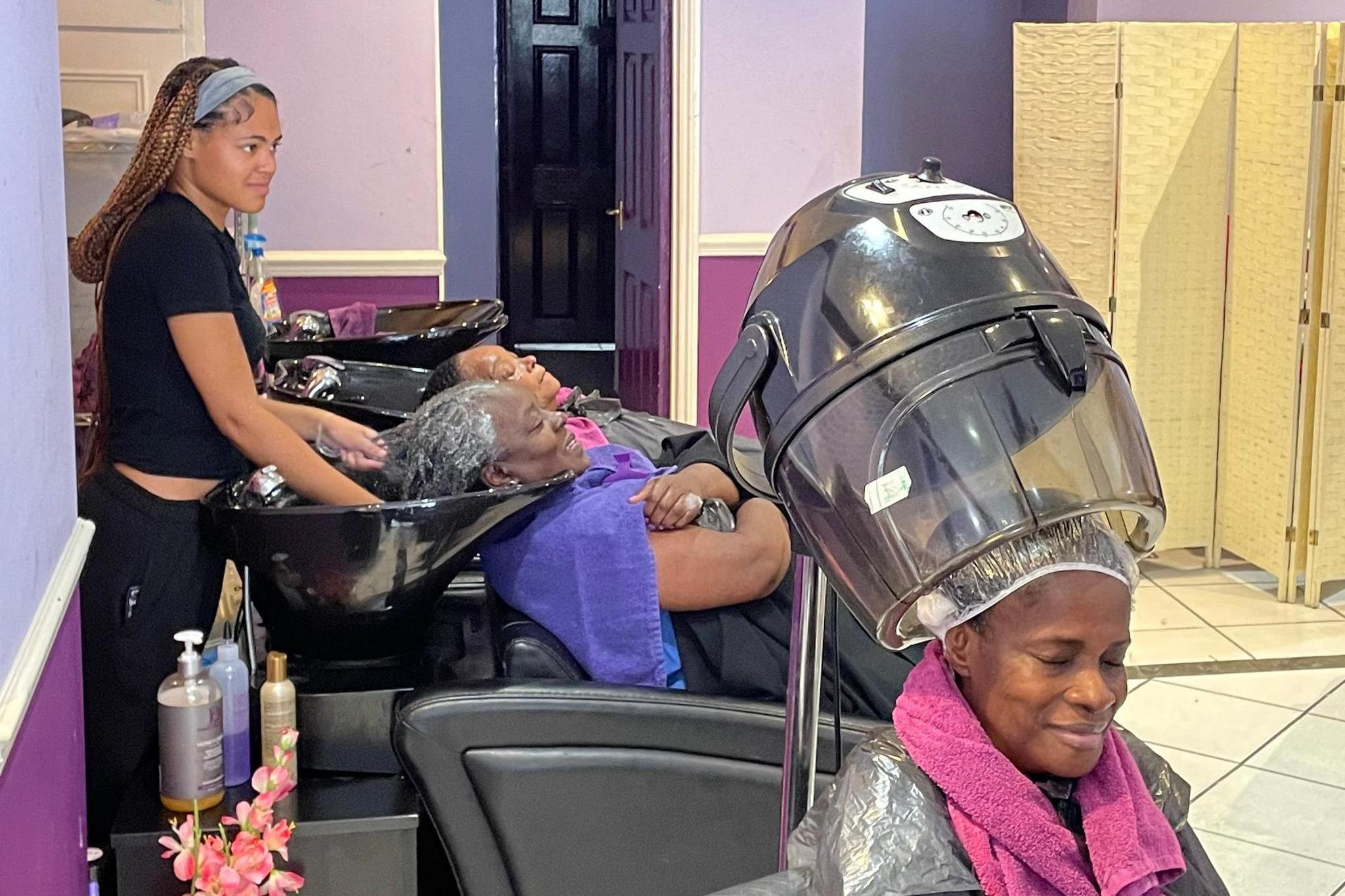 Inside Pride Salon, a client sits beneath a large hairdressing drying hood, while in the background junior assistant Jahmelia Hoilett shampoos another client’s hair at the wash station.