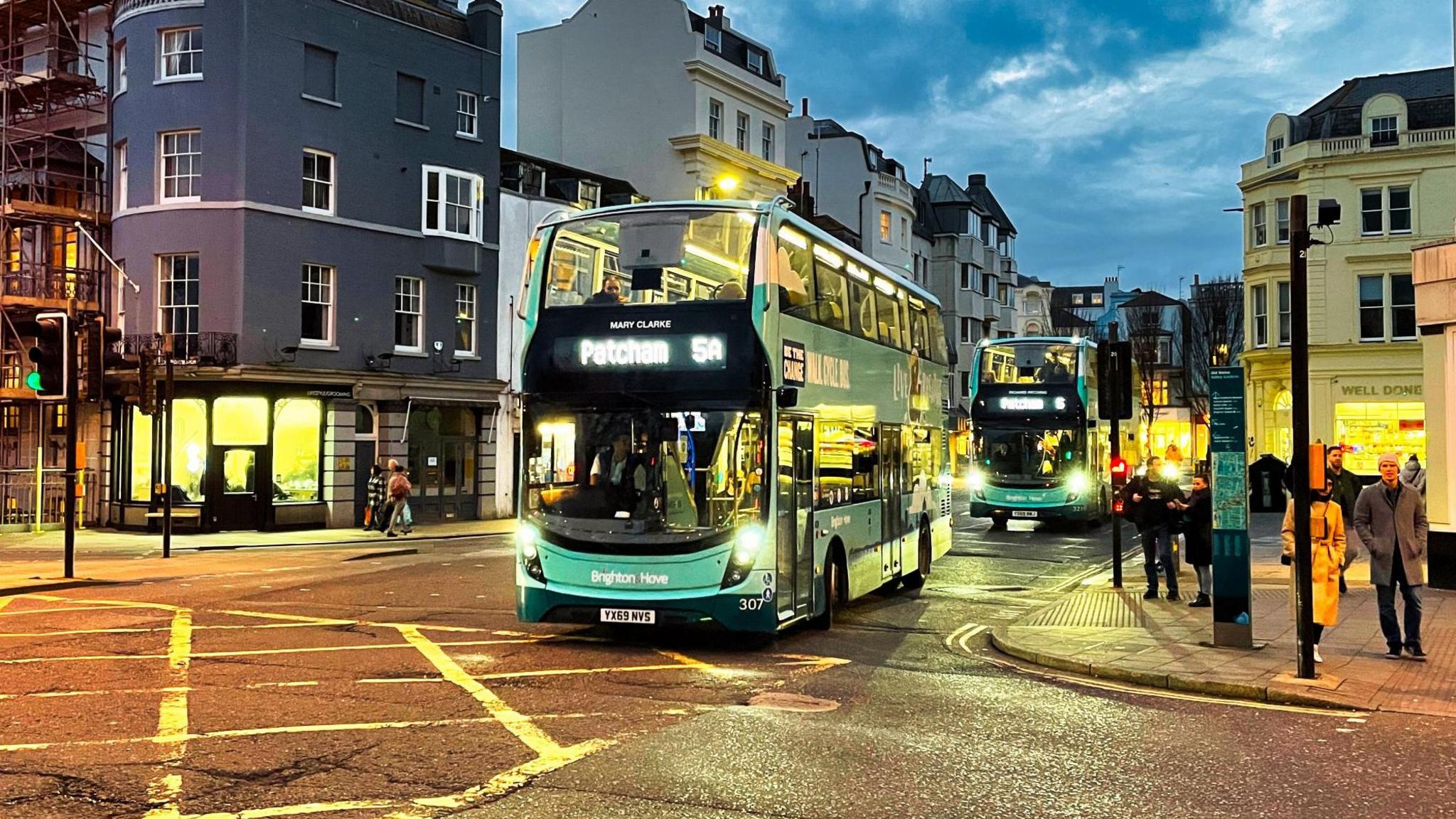 A baby blue bus rides through a city at dusk