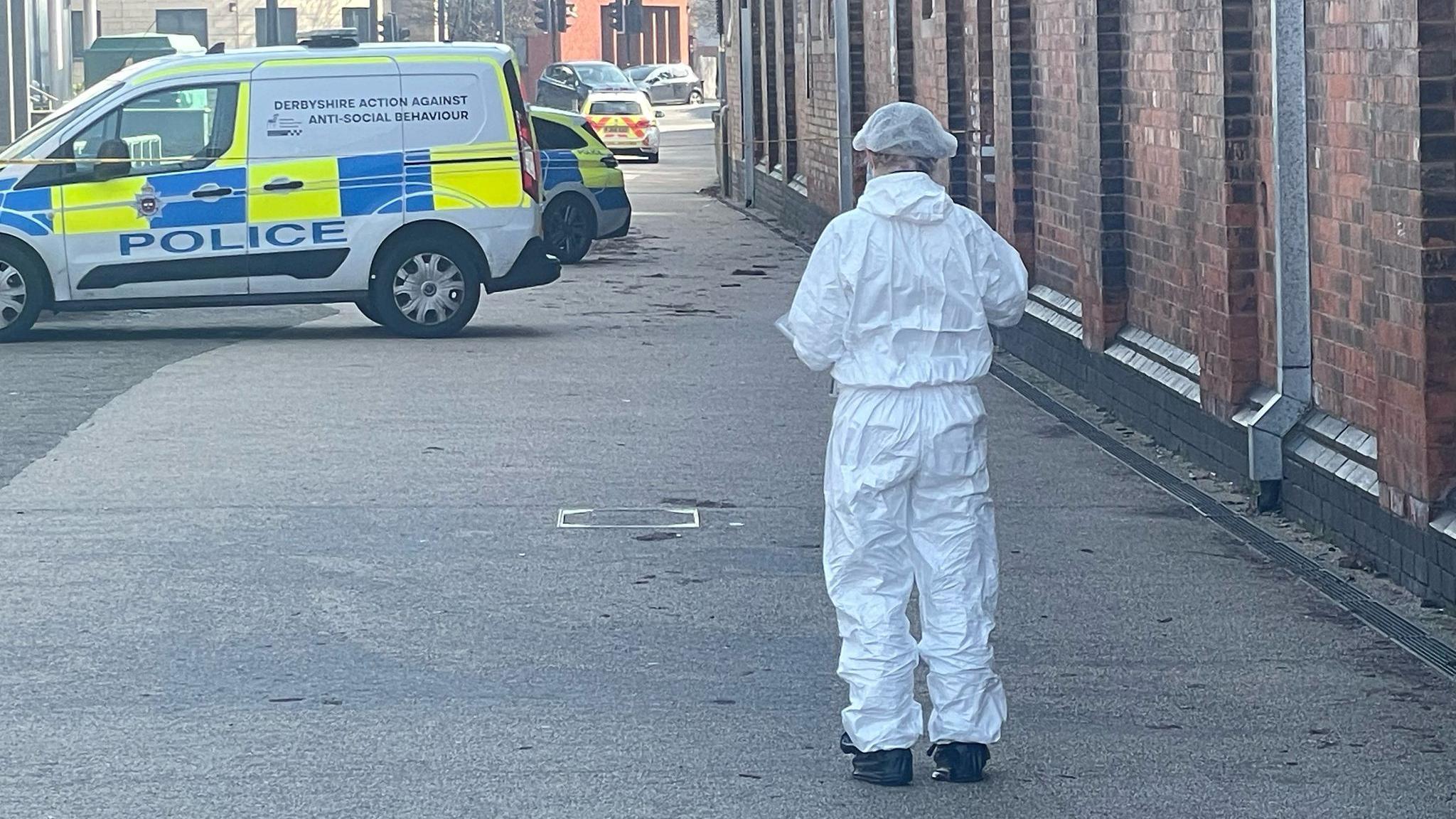 A man wearing a white forensics suit is stood in a walkway with his back to the camera. Ahead of him is a police van and behind the van, two police cars can be seen. 