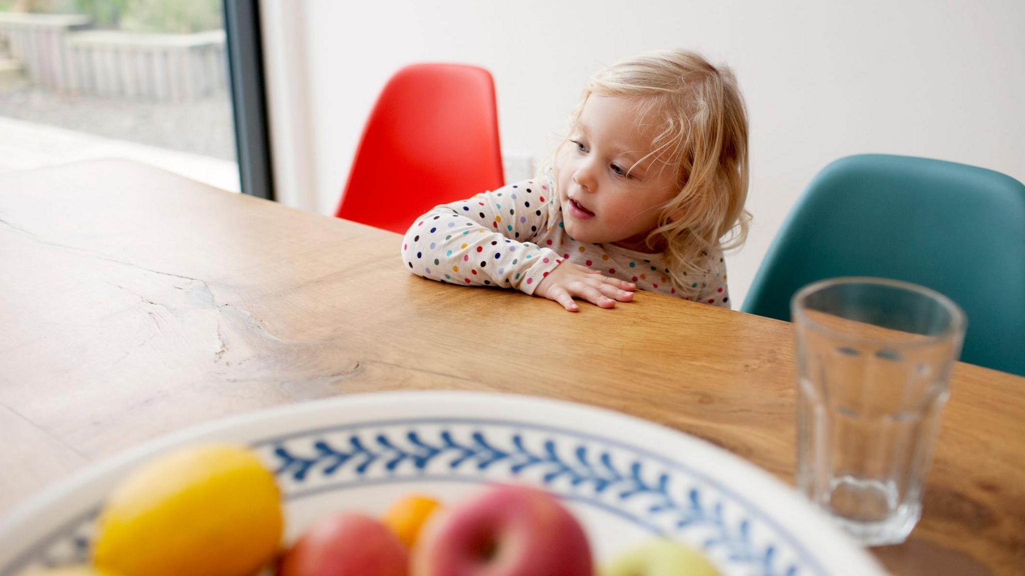 A child with blonde hair standing in front of a brown oak table with a glass and a bowl of fruit on it. 