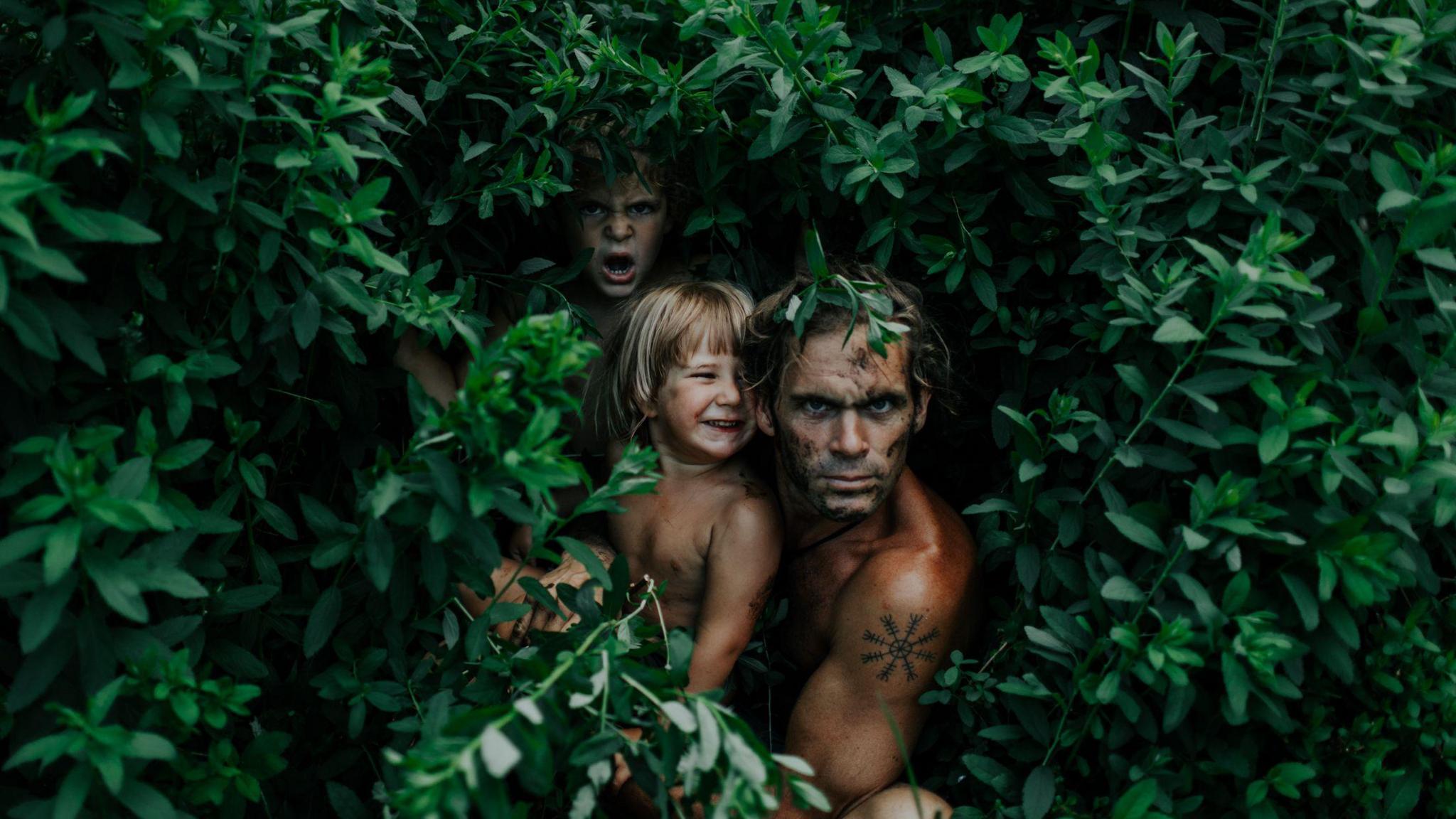 Nik Payne, who is topless, peers from inside a leafy bush with his young sons on his knee
