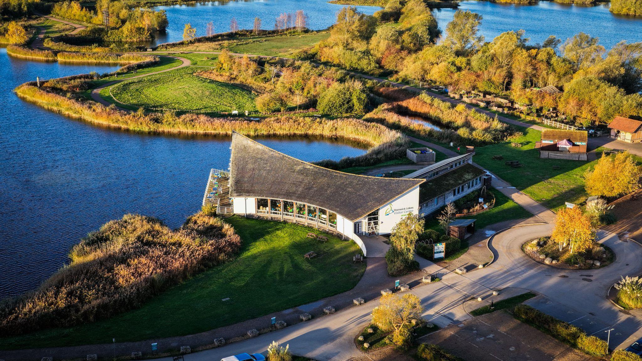 A drone shot of the main visitors' centre of Stanwick Lakes, surrounded by countryside and waterways. It is a white building with a sloped roof. It is autumn and the trees are all turning brown.