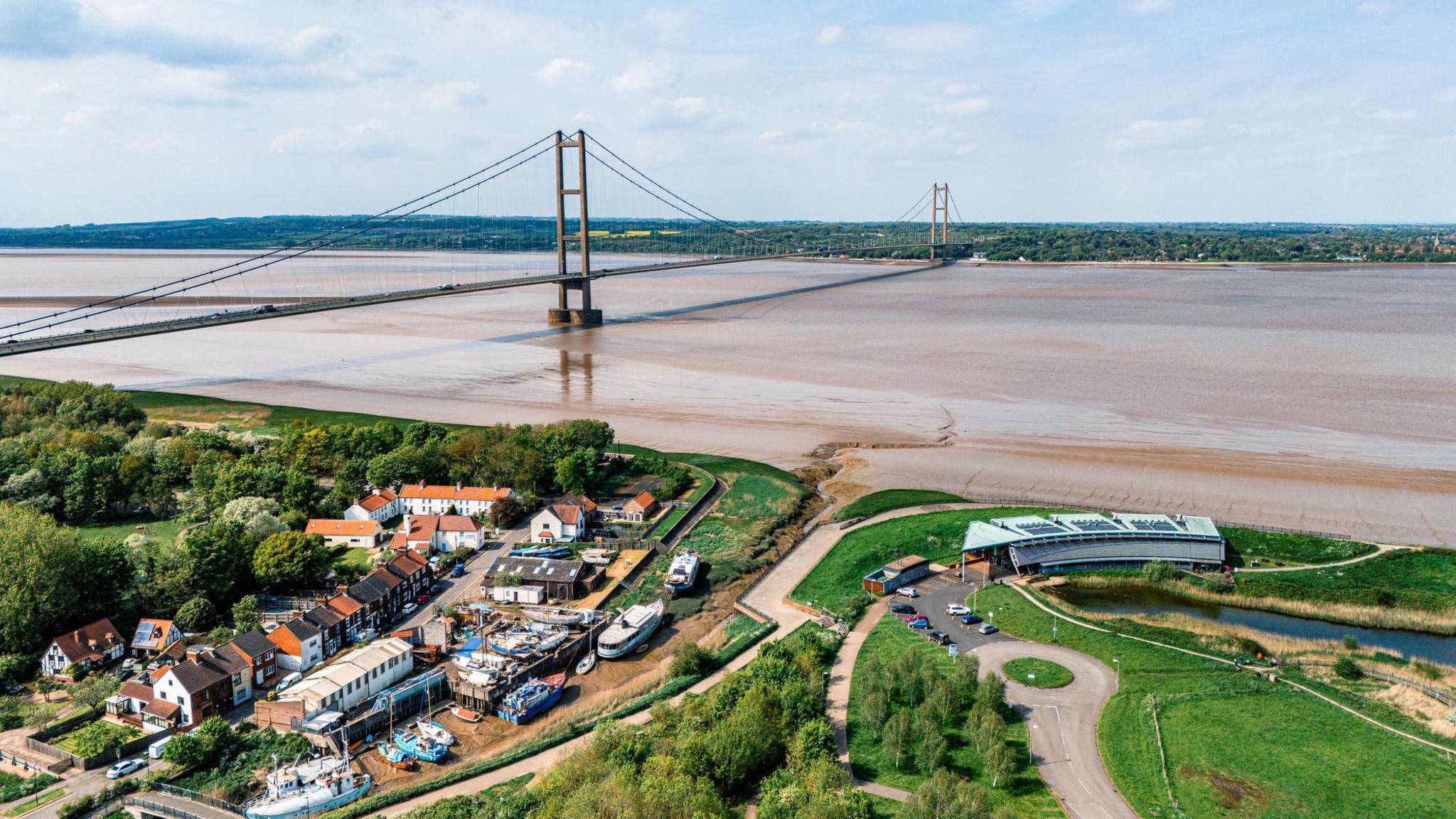 The Humber Bridge from distance. The structure is standing over a low tide with a small community in Barton on the edge of the water. It is surrounded by plenty of trees and green land.