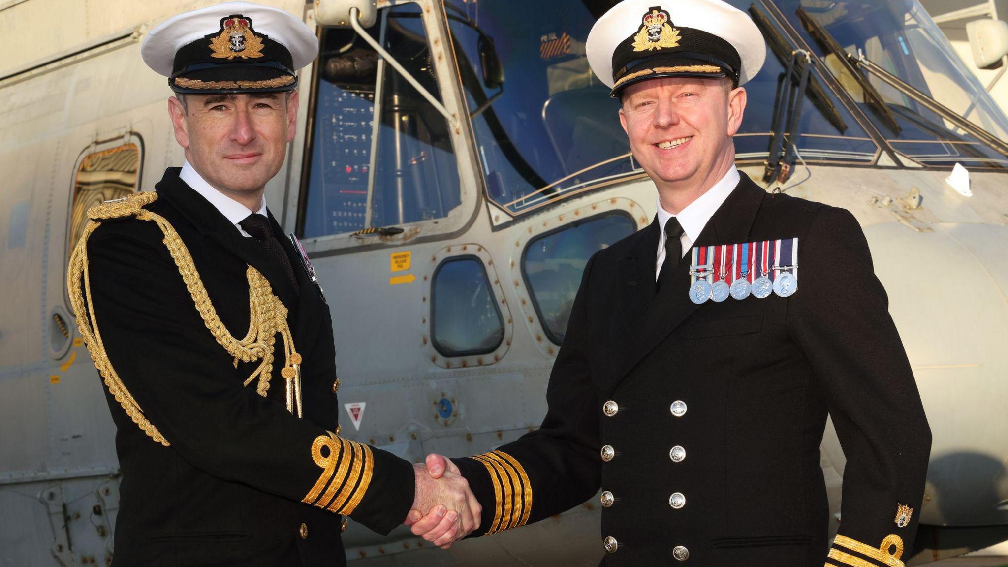 Capt Stuart Irwin hands over command of RNAS Culdrose to Capt James Hall. They are both wearing senior naval dress uniforms, with medals on, shaking hands in front of a grey naval helicopter.