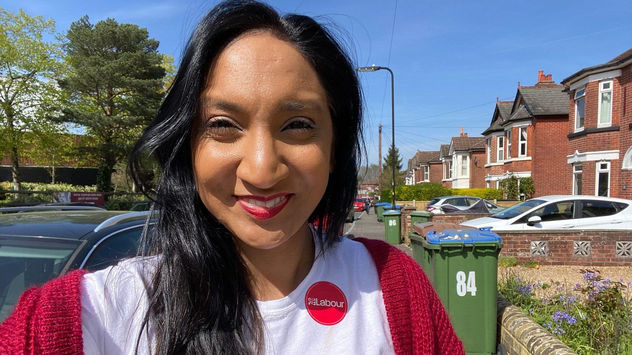 Satvir Kaur is standing in a rediential street on a sunny day, she is taking a selfie, wearing a white t shirt with a circular red Labour sticker on the left hand side and a red knitted cardigan over her shoulders.