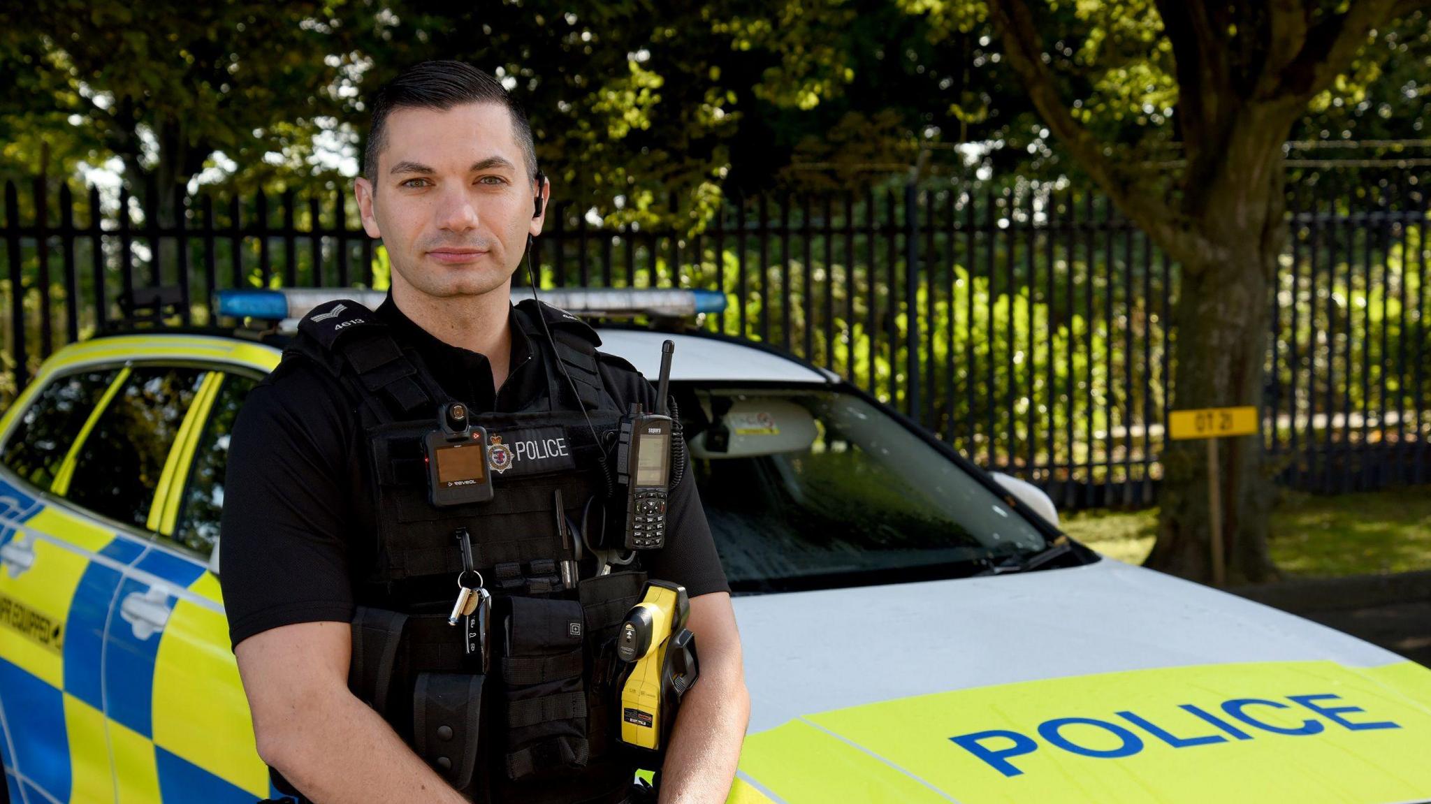 Sgt Luke O’Connell, from Avon and Somerset Police, stands facing the camera. He is in his full police uniform and is next to a police vehicle with yellow and blue livery