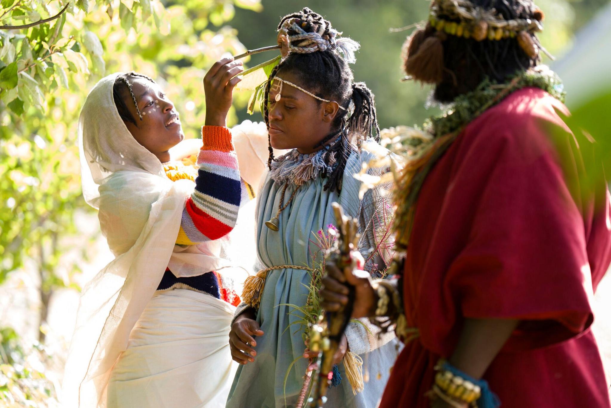 Handmaiden 'Asnat' wears a white shawl and dress with a colourful striped jumper underneatch and styles 'Queen Nandi''s hair. She wears a blue dress and stands next to 'King Atehene' who is dressed in red at their campsite in woods near Jedburgh. 