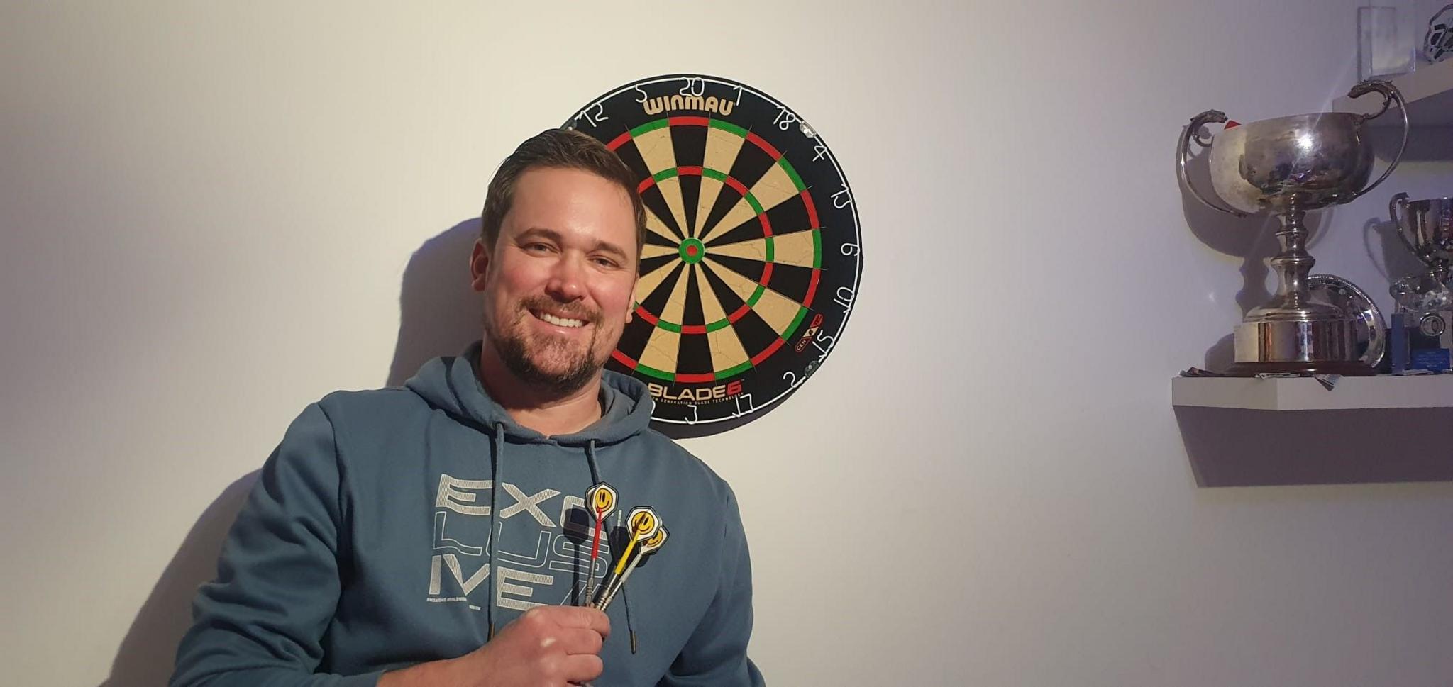 Ian Driver is holding a set of darts and is standing in front of a board and a shelf holding his trophies