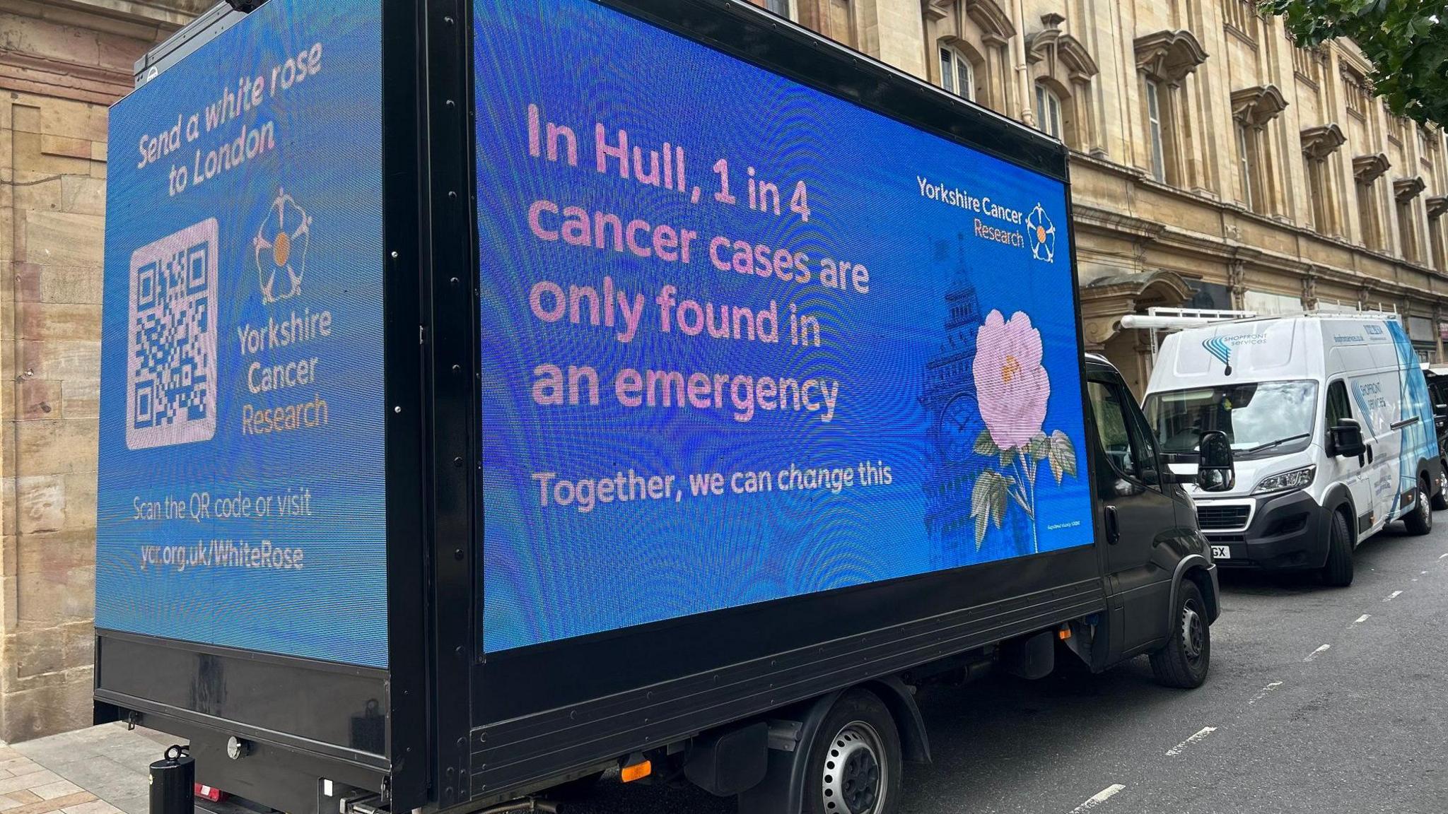 A blue van displaying digital billboard for Yorkshire Cancer Research is parked outside a grand, stone-built city hall. Messages read: "Send a white rose to London", and "In Hull, one in four cancer cases are only found in an emergency - together we can change this".