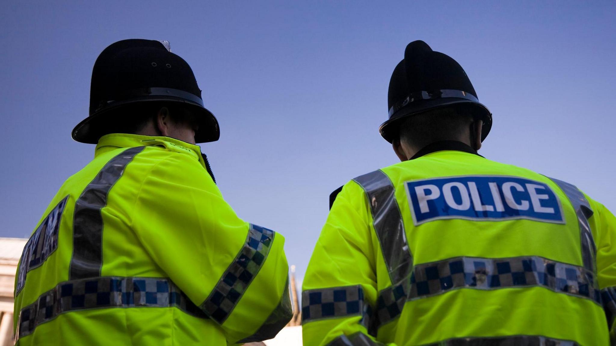 Two police officers facing away from the camera. Both are wearing yellow hi-vis jackets with a blue and white sign reading "Police" on the back.