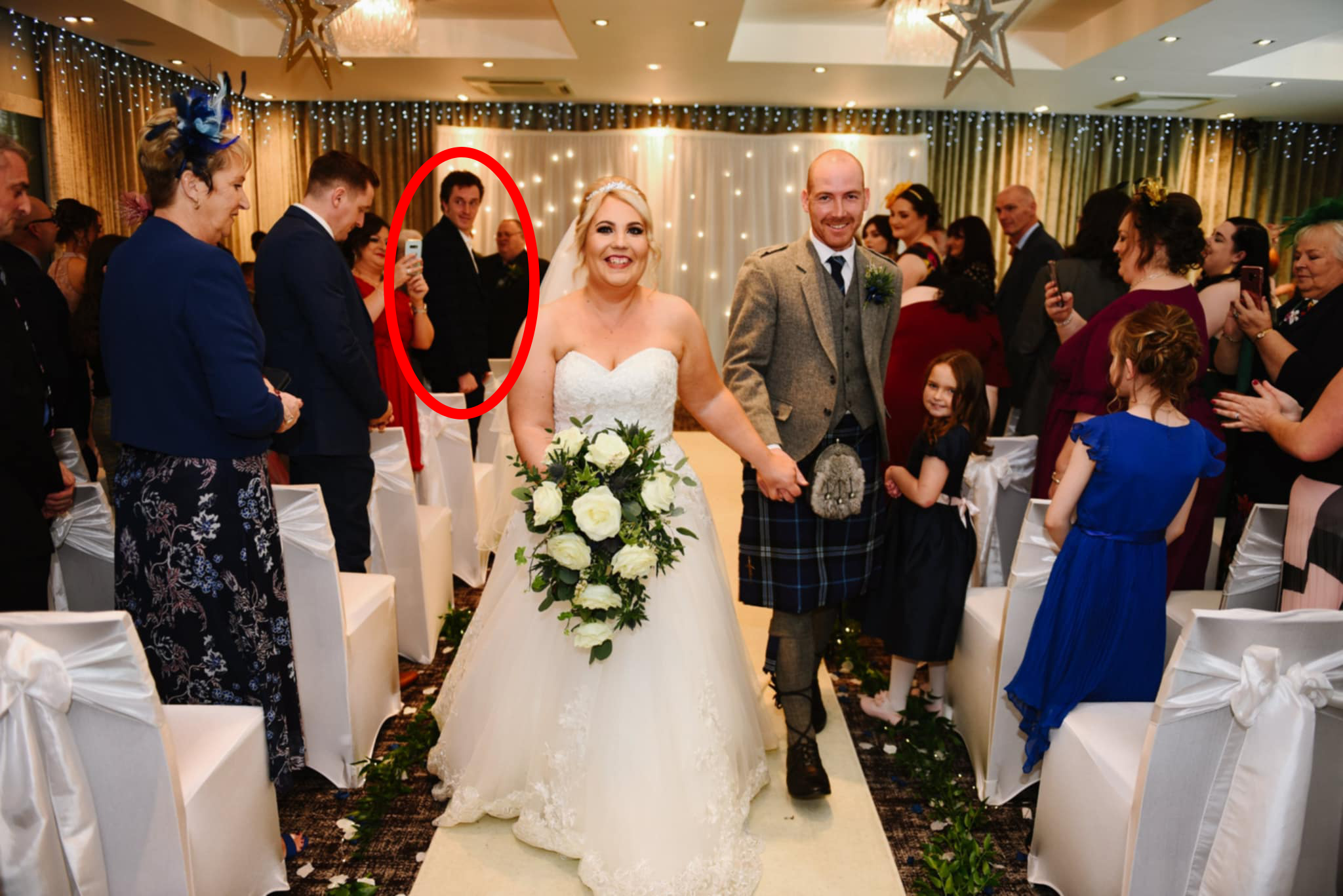 A happy bride and groom walking back up the aisle after their wedding, with beaming smiles on their faces. Many of the guests are applauding them. Andrew Hillhouse can be seen in the background as one of the attendees, with a red ring overlain on the image to draw the eye to him.