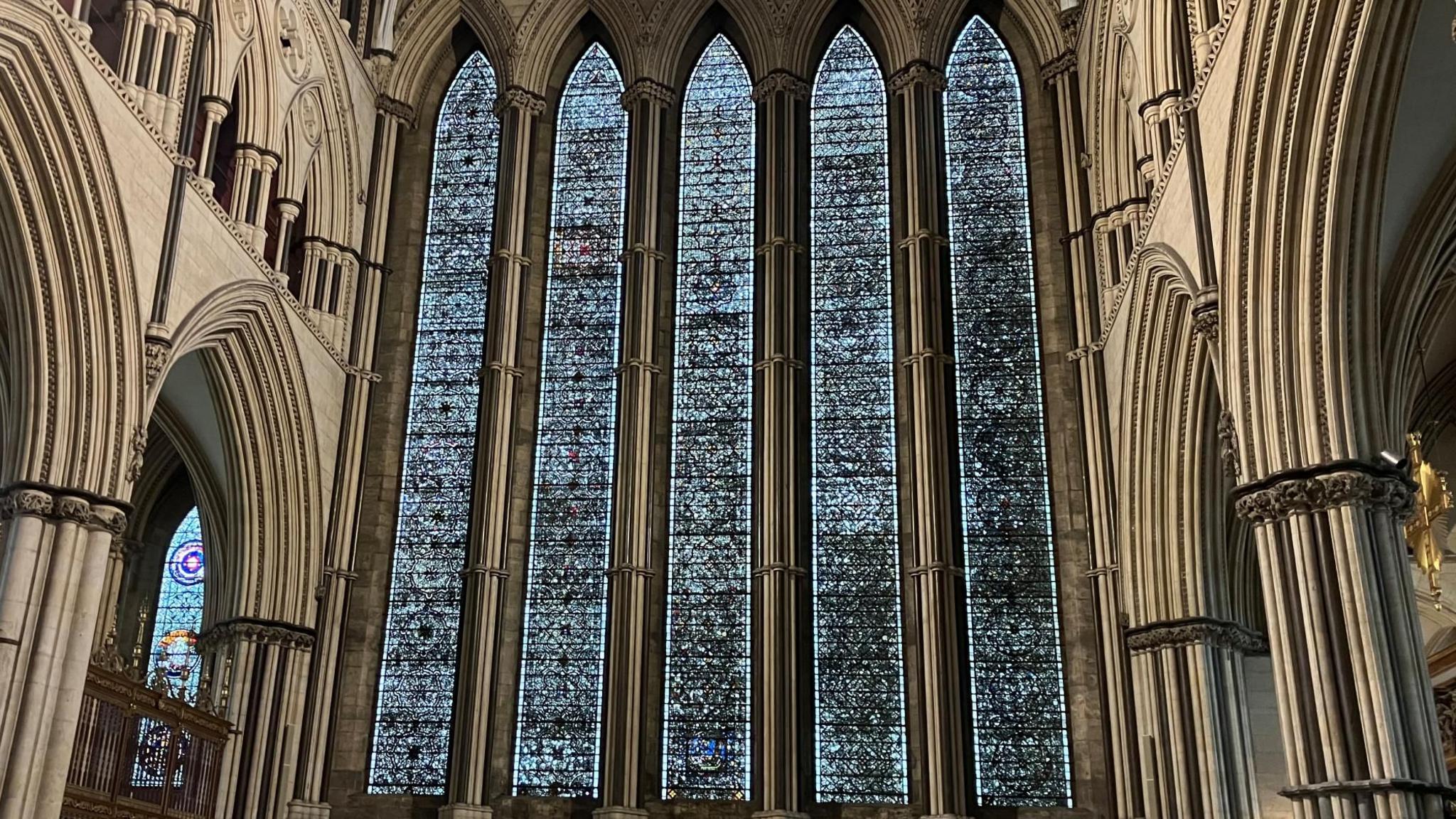 An interior view of a large, five lancet, stained glass window, set in a stone cathedral.