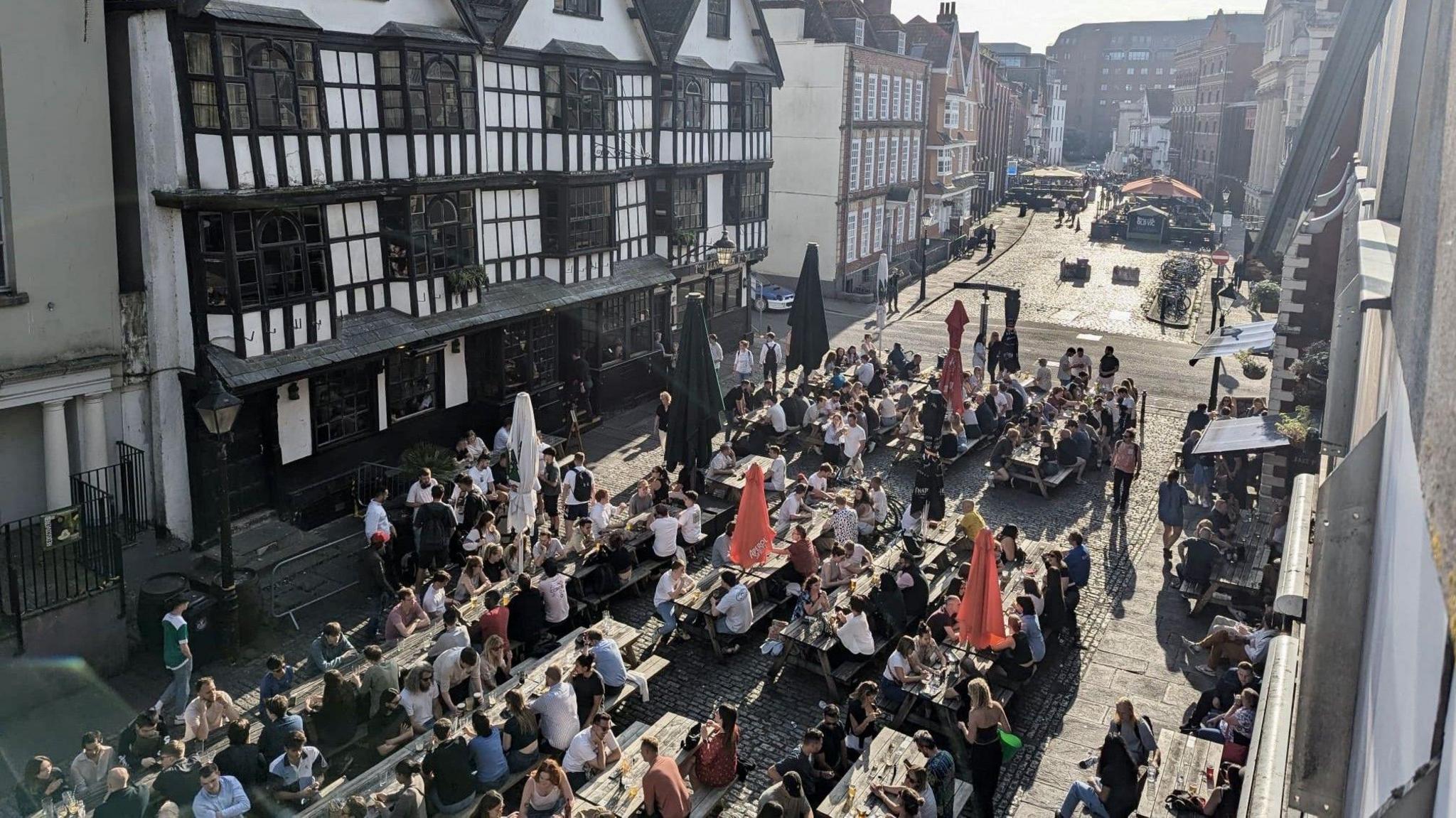 King Street in central Bristol is visible in a photograph taken from a balcony overhead. Dozens of people are sitting at wooden tables in the street and in the background the dark wood and white paint frontage of the Llandoger Trow pub is visible.