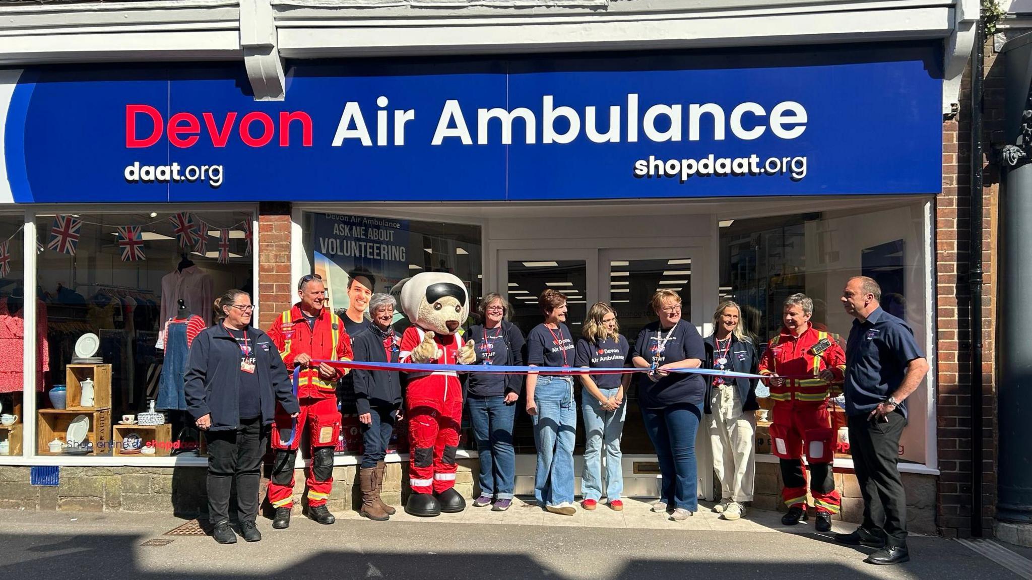 A ribbon-cutting ceremony in front of a store a newly opened Devon Air Ambulance charity shop. There are 11 people stood outside the shop all wearing company uniform. One is dressed as a mascot.